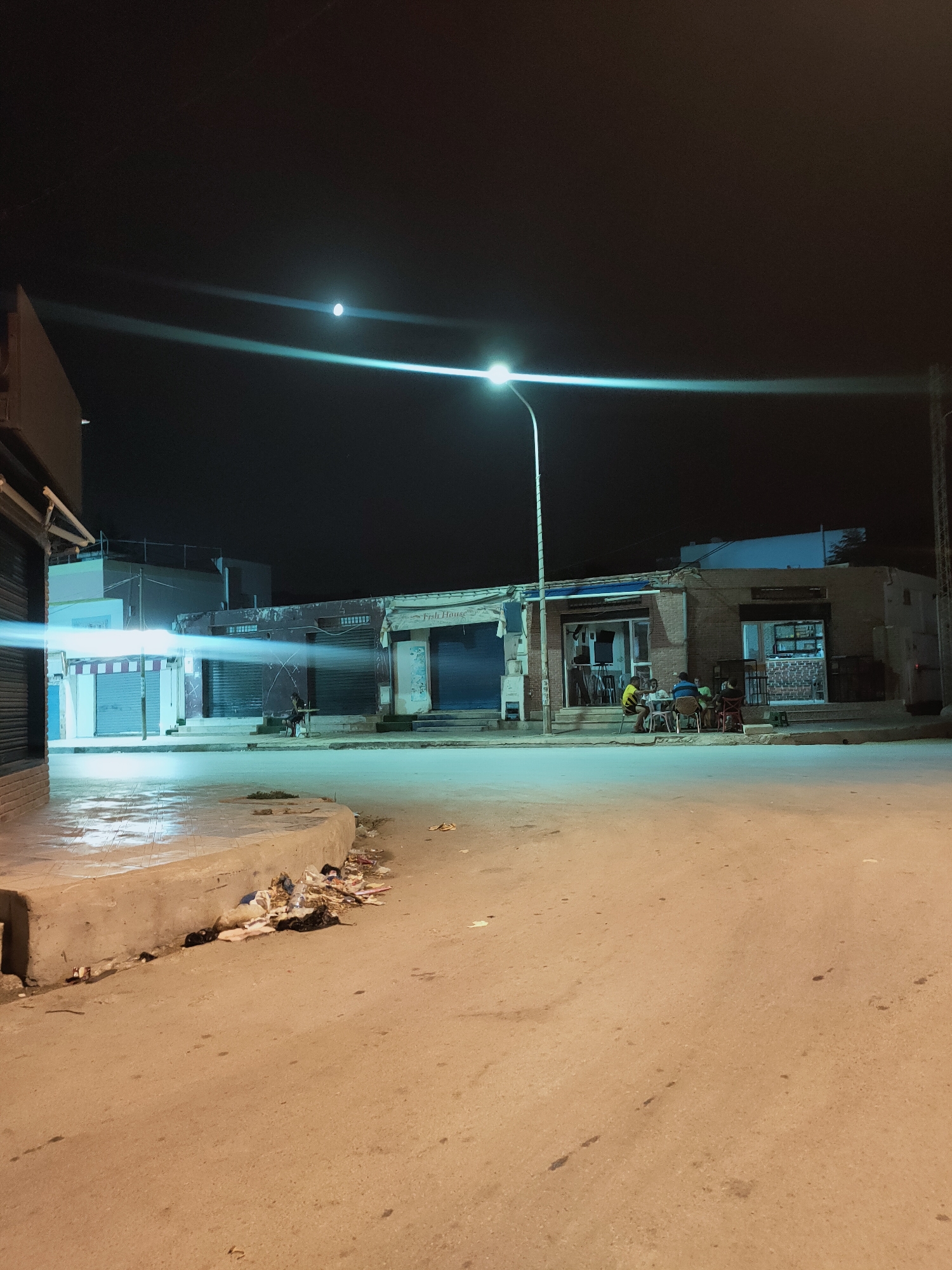 This nighttime street scene, captured in Ariana, Tunisia, depicts a moderately lit urban environment. The foreground is dominated by a paved road surface with scattered debris, including plastic bottles and bags, concentrated near a raised concrete curb on the left. Beyond the road, a sidewalk runs in front of several low-rise buildings.
To the left, a building with shuttered storefronts is partially visible, illuminated by a bright, possibly street lamp-induced, light beam that streaks horizontally across the image. Further along the sidewalk, a series of shops with closed metal shutters are evident. One establishment features an open doorway with interior lighting and what appears to be a sign reading "Fish House."
In front of the "Fish House" building, a group of four individuals are seated at a table, engaged in conversation or dining. One person is wearing a yellow and black striped shirt. Another building to the right also has an open storefront with interior lighting, displaying shelves with items.
A tall street lamp stands on the left side of the street, its light casting a bright beam and contributing to the overall illumination of the scene. The night sky above is dark, with a bright circular object, likely the moon, visible in the upper left quadrant, accompanied by light streaks, possibly lens flares. The architectural style of the buildings suggests a typical North African or Mediterranean urban setting.