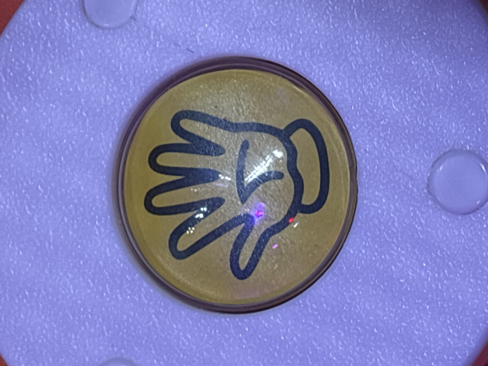 A close-up, top-down view shows a yellow, circular button with a black outline of an open hand. The hand is depicted with the palm facing forward and fingers spread. The button is set against a textured, lavender-colored plastic surface. Two small, white, circular protrusions are visible on the lavender surface, one to the left and one to the right of the button, partially cropped out of the frame. There are reflections of light on the button, including a prominent blue and pink glint on the palm of the hand. No specific location in Dubai, United Arab Emirates, can be determined from this image.