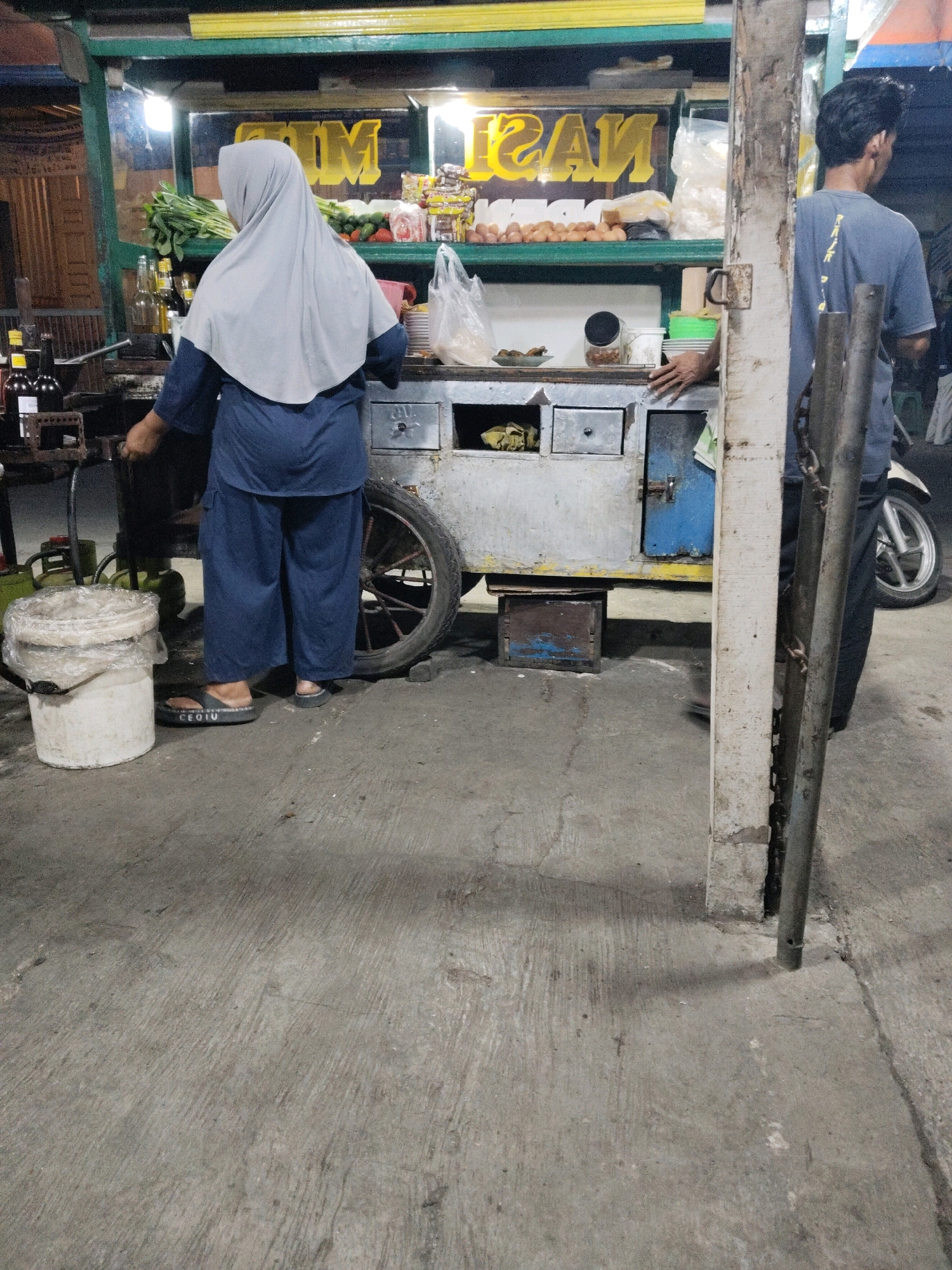 The image depicts a street food stall in Subang, Indonesia, at night. A woman wearing a grey hijab and blue attire stands behind the counter, facing away from the camera. In front of her is a bucket wrapped in plastic. To her right, a man in a blue t-shirt and dark pants stands near a motorcycle. The stall itself is a cart with a metal body, featuring shelves stocked with various food items including eggs, vegetables, and packaged goods. A large wheel is attached to the side of the cart. The ground is paved with concrete, showing some wear and tear. The lighting suggests an outdoor, dimly lit environment.
