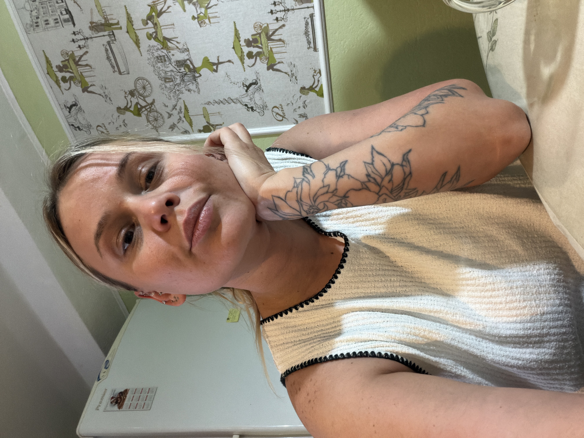 A woman with fair skin and blonde hair is posing for a selfie in an indoor environment. She has a floral tattoo on her left arm. She is wearing a white textured top with black trim. Her hand is near her face, and she has multiple piercings in her left ear. Behind her is a light green wall and a refrigerator. Above her is a wall covering with a patterned design. The exact location in Sarapul, Russia, is not evident from the image.