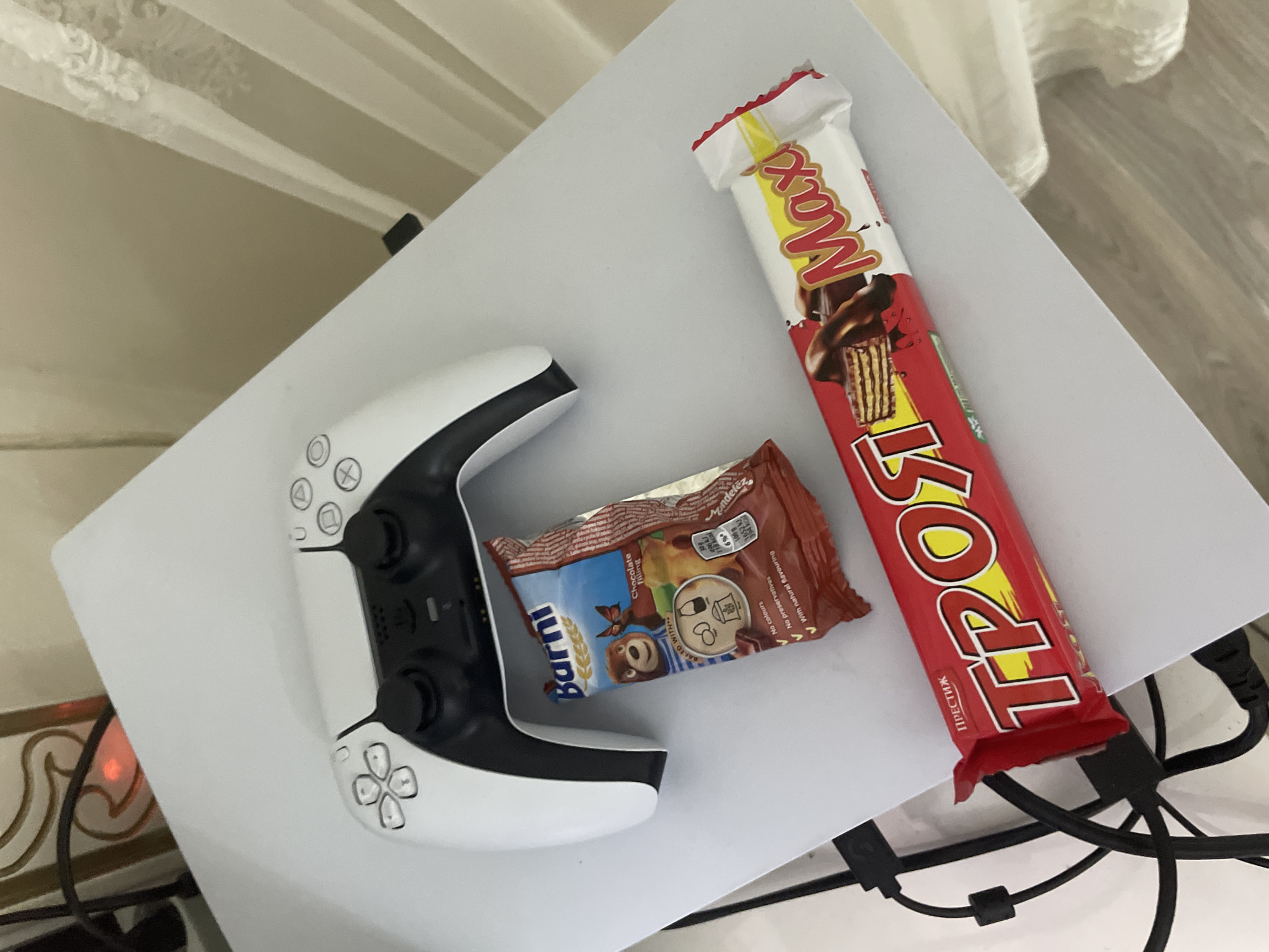 A high-angle, close-up shot displays a gaming setup with snacks on a white, flat surface, likely a PlayStation 5 console, situated indoors in Ihtiman, Bulgaria.
On the left, a white and black PlayStation 5 DualSense controller is prominently featured, with its buttons, including the directional pad and face buttons (circle, cross, square, triangle), clearly visible. A subtle red light emanates from the lower left, possibly indicating the console's status or the controller's charging state.
Adjacent to the controller are two snack items. A small, brown packet of "Barni" with "Chocolate Filling" is visible, showcasing a cartoon bear and butterfly. The packaging states "Baked With," "No colours," "No preservatives," and "With natural flavouring," along with nutritional information from "Mondelēz."
To the right, a larger red and yellow packaged chocolate bar stands out. The wrapper features the words "Max" and "TPOPI" in bold letters, and the Cyrillic text "ПРЕСТИЖ" (PRESTIGE) is also discernible. The image on the packaging depicts a chocolate-covered wafer bar.
In the background, light-colored wooden flooring and drapes or curtains suggest a domestic setting. Black cables with a ferrite bead are visible in the lower right, disappearing behind the white surface. The arrangement implies a leisurely activity, such as gaming, with snacks at hand. The visible Cyrillic text and snack brands are consistent with the provided location context of Bulgaria.