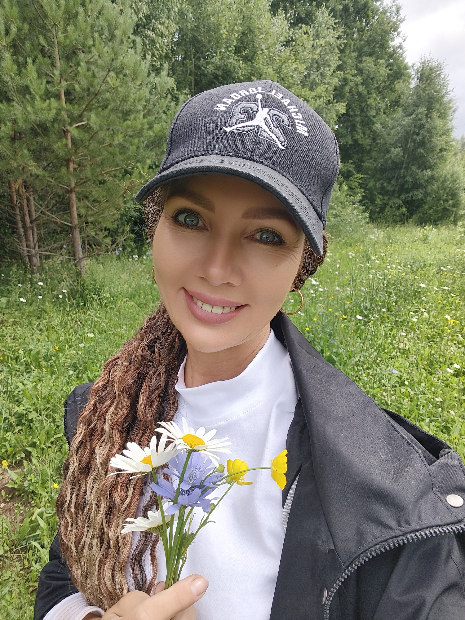In the city of Noviy, Russia, a woman is pictured in a selfie from the chest up. She is wearing a black baseball cap with a white logo on it, a white turtleneck shirt, and a black jacket. Her long, wavy, brown hair is visible on the left side of her face, and she is holding a small bouquet of white and blue flowers in her right hand. She has a bright smile and blue eyes. The background consists of a grassy field with scattered wildflowers and trees. The lighting suggests it is daytime.