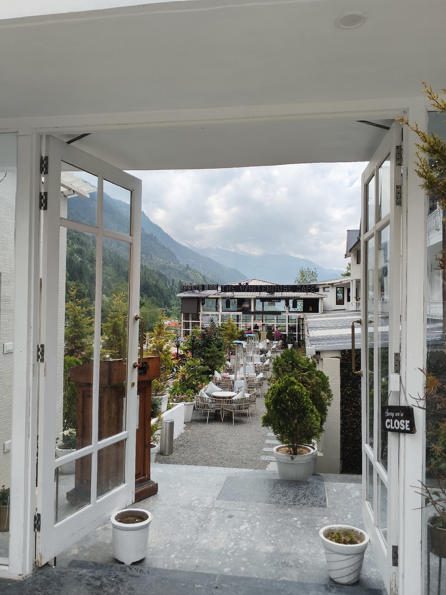 This media file depicts an outdoor seating area of a establishment, possibly a restaurant or cafe, with a stunning mountainous backdrop. The perspective is from an open doorway, looking out onto a patio furnished with numerous round tables and chairs, all made of woven material with light-colored cushions. Several large, cylindrical heaters stand among the tables, suggesting the possibility of cooler weather or evening use.
The setting appears to be in a hilly or mountainous region, evidenced by the lush green slopes covered in trees that rise in the distance. Snow-capped peaks are visible further back, indicating a high-altitude location. The architecture of the buildings in the background is modern, with flat roofs and balconies. One building has a sign that reads "SPORTS BAR".
The foreground shows a grey, tiled floor with several white planters, two of which contain small, potted plants. The doorway itself is white, with glass panels that offer a clear view of the exterior. On the right-hand door, a small, black sign hangs, displaying white text that reads "Sorry we're CLOSE".
The weather appears to be overcast, with a cloudy sky that diffuses the light, suggesting it could be morning or late afternoon. The overall mood is peaceful and inviting, despite the "closed" sign, conveying a sense of a serene escape within a natural landscape. No people are visible in the scene. The location is identified as Barheta, India.