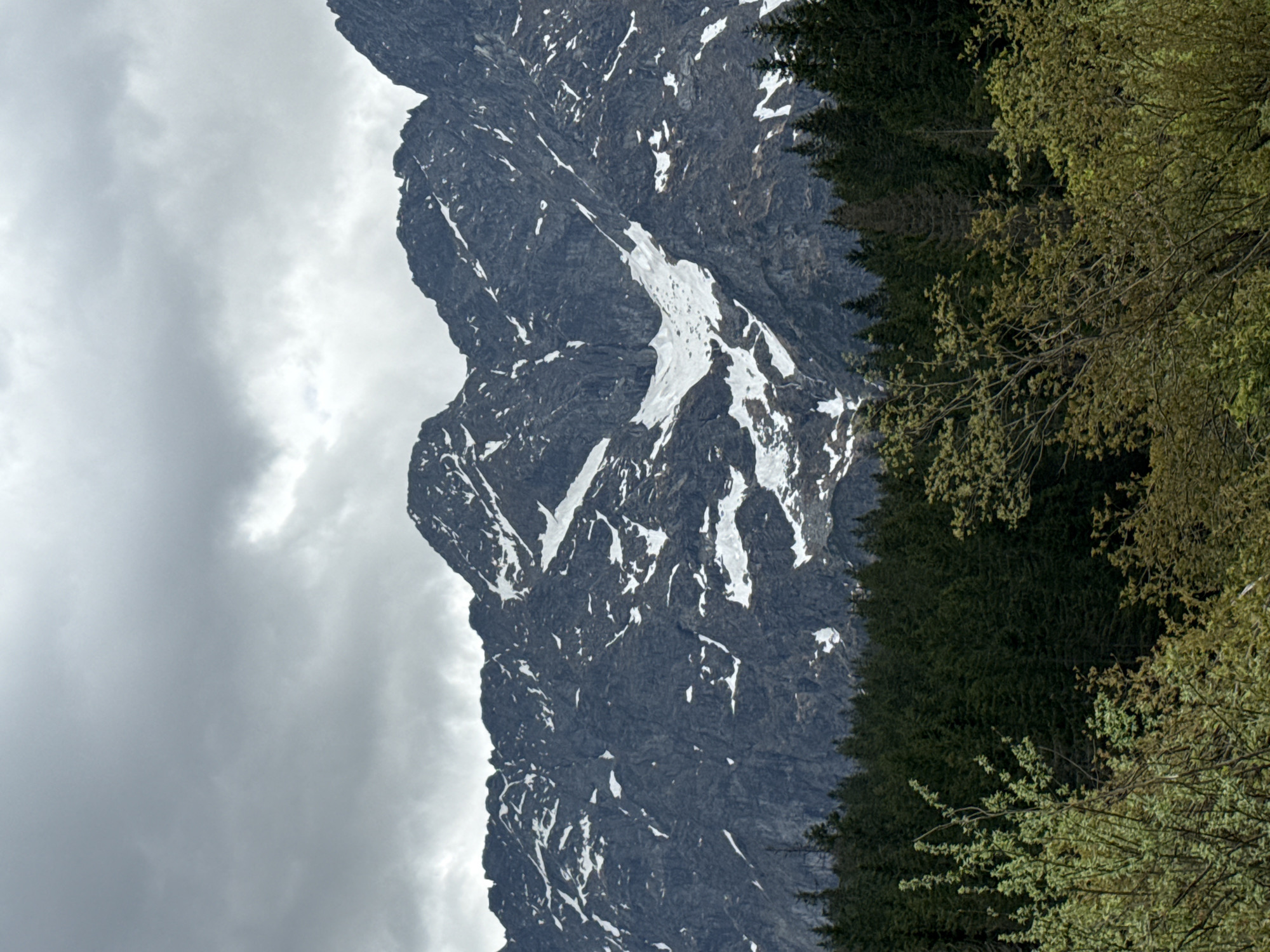 The image depicts a mountain with snow patches on its rocky face. To the right of the mountain are dense, evergreen trees, and some deciduous trees with budding leaves. The sky is overcast with heavy clouds. The scene is not recognizable as being located in Brzegi, Poland. The image is not identifiable in Brzegi, Poland.
