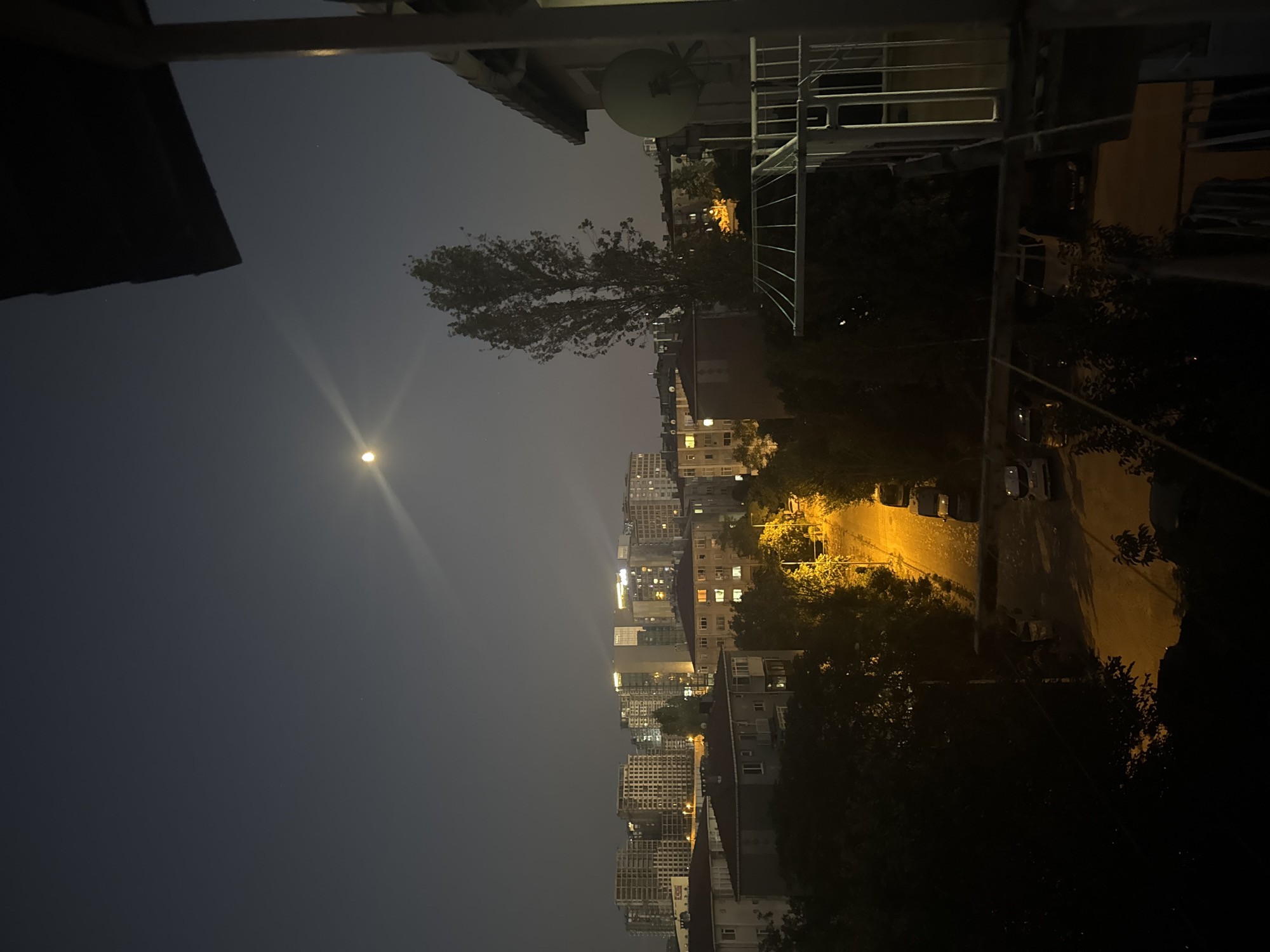 A nighttime, slightly hazy aerial view shows a residential street in Gungoren, Turkey. A bright moon hangs in the dark sky, with a visible light flare. The street below is illuminated by orange streetlights, casting shadows on the asphalt. Several cars are parked along the side of the road. Dense foliage, including trees and bushes, lines the street, creating a dark, natural border. In the background, a cluster of multi-story apartment buildings and construction sites are visible, their windows lit up, indicating inhabitation. The foreground features a balcony railing and the edge of a building's roof.