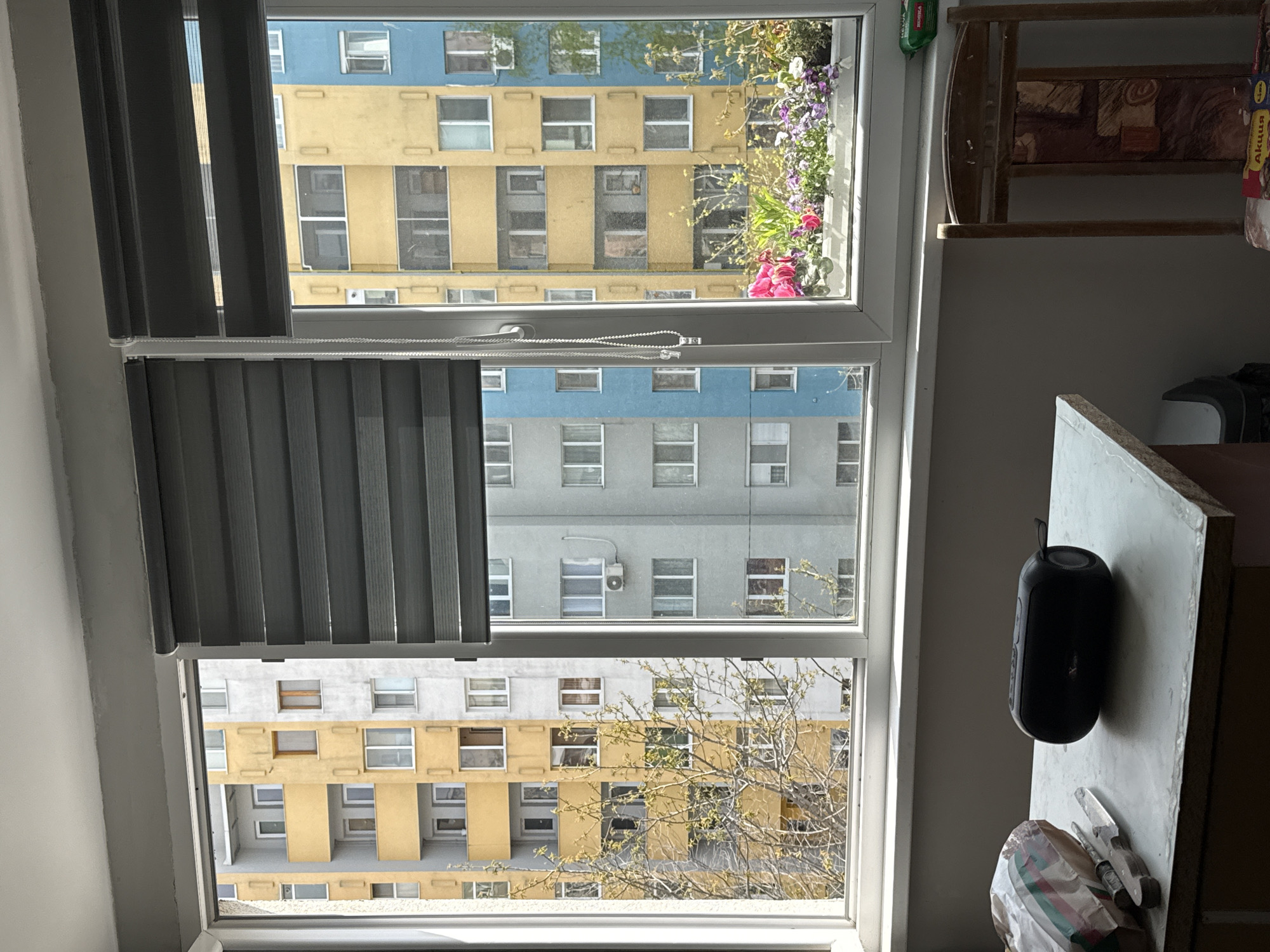 A window with gray blinds offers a view of an apartment building, located in Stara Zagora, Bulgaria. The building is painted in alternating sections of yellow and blue, and another is gray. The window is trimmed in white, and a small potted plant with pink and purple flowers sits on the window sill. To the right of the window, there is a shelf and a white countertop. A black speaker is visible on the countertop. A plastic bag is also on the countertop.