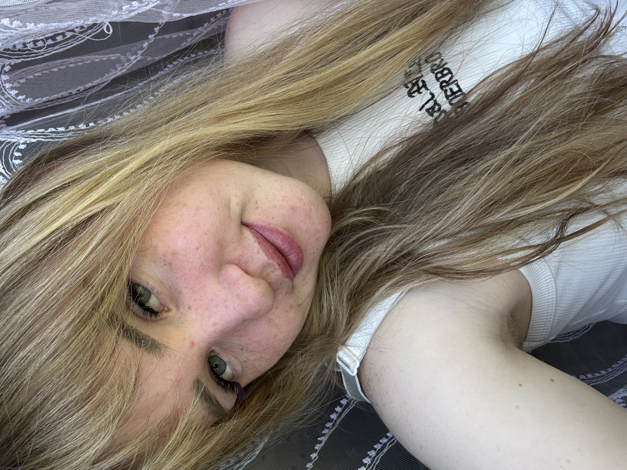 A young woman with fair skin and light brown hair lies on a patterned bed covering. She is wearing a white top with black lettering. The location is Марковский, Russia.