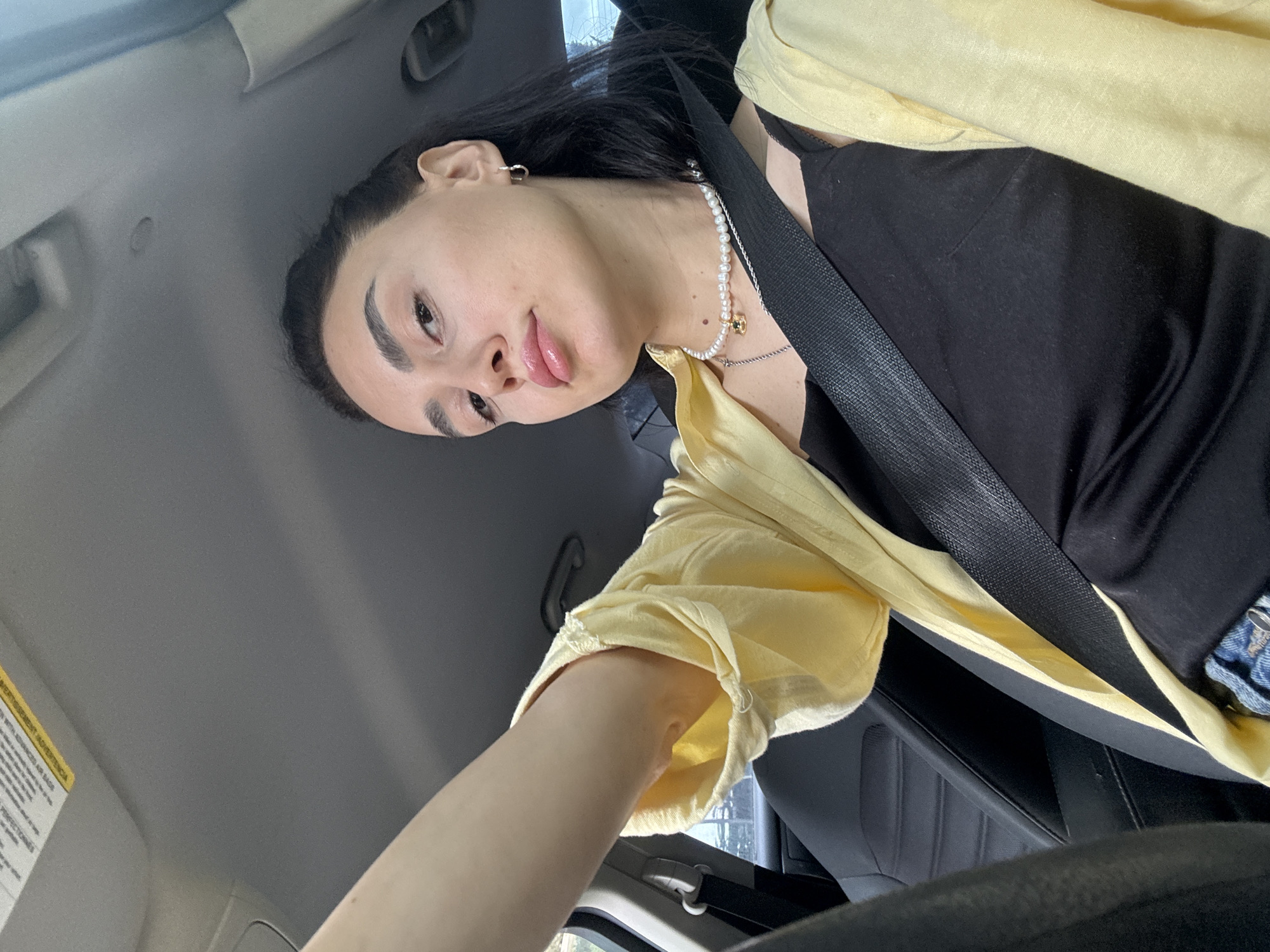 A person is seated in the driver's seat of a car, wearing a black top, a yellow blouse, a pearl necklace, and a seatbelt. The car's interior is visible, including the dashboard and steering wheel. The location is likely Tbilisi, Georgia, although this cannot be definitively confirmed from the image alone. No specific actions or interactions are depicted beyond the person posing for the photograph.
