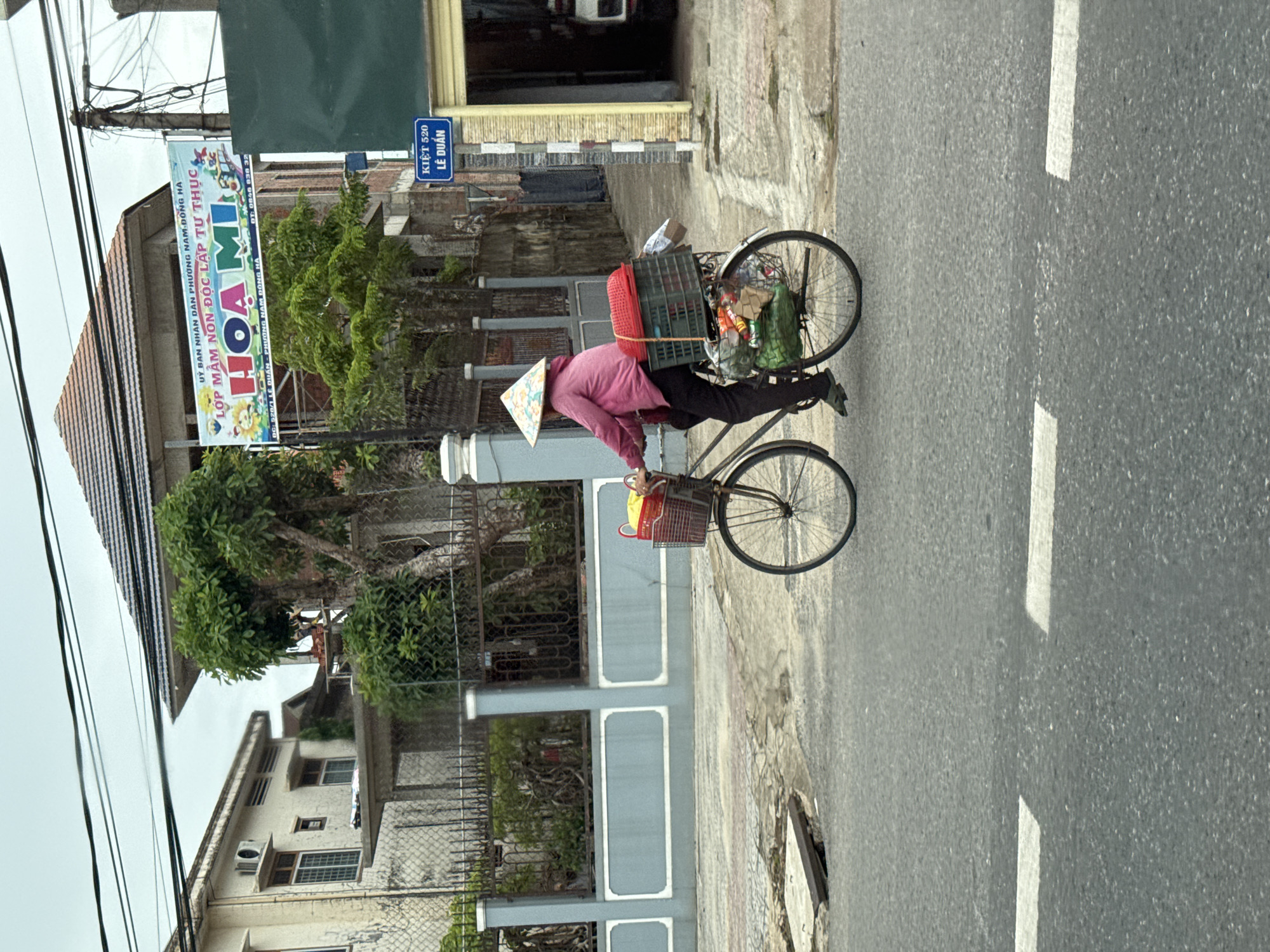 In Nam Đông Hà Ward, Vietnam, a person wearing a conical hat and a pink jacket rides a bicycle loaded with goods. The bicycle has two baskets attached, one in the front and one in the back. The rear basket is filled with green bags and other items, topped by a red woven container. The person is dressed in dark pants and green shoes.
The scene unfolds on a paved road with white lane markings. To the left of the road, a light blue wall with white decorative elements runs along a property, bordered by a metal fence. Behind the fence, a building with multiple windows and an air conditioning unit is visible, along with a large green tree.
In the background, a building with a dark-tiled roof is under construction or renovation, with scaffolding visible. A banner advertising "HOA MI" preschool hangs from the building. A blue street sign indicates "KIỆT 520 LÊ DUẨN". To the right, a building with a yellow facade and a garage-like opening is present. The sky is overcast.