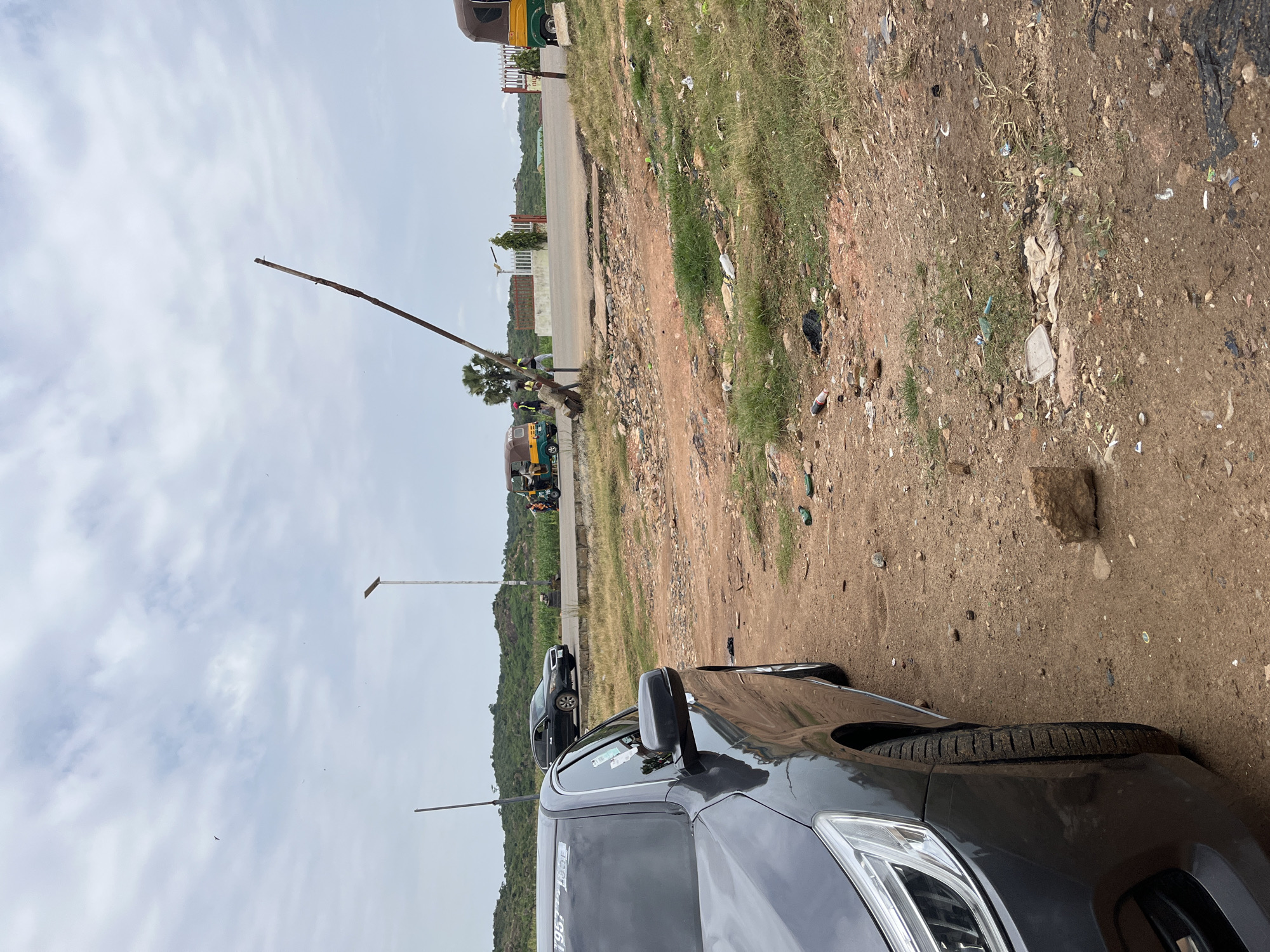 The image depicts a roadside scene in Jos, Nigeria. In the foreground, the left side of a dark grey sedan is visible, parked on a dirt and gravel surface. The car's tire and side are in focus. The background reveals a slightly uneven asphalt road, with several auto-rickshaws (keke napep) and a few people. One auto-rickshaw is yellow and green, with passengers. Another, also yellow and green, is partially visible on the right. A tall, thin metal pole, angled upwards, stands near the road. Beyond the road, rolling hills with sparse vegetation are visible under a cloudy sky. The ground beside the road is littered with trash, including plastic bottles and bags. A solar-powered street light is present in the middle ground.