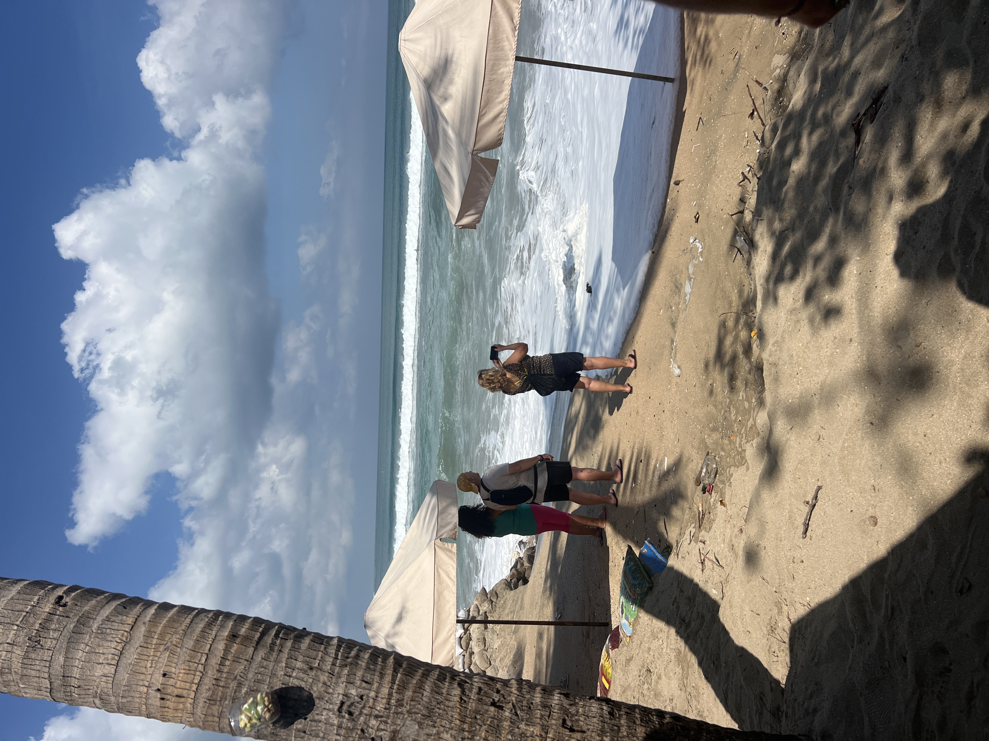 A candid, eye-level shot captures a trio of individuals standing on a sandy beach in Kuta, Indonesia, facing a turbulent ocean. The water is a vibrant turquoise, with white frothy waves crashing ashore, creating a foamy edge along the sand. In the distance, the ocean stretches out to meet a bright blue sky dotted with fluffy white clouds.
The people are positioned from left to right: a woman with long, dark curly hair wearing a green top and pink shorts; a man wearing a light-colored t-shirt, dark shorts, and a yellow cap, with a black backpack slung over his shoulder; and a woman with blonde hair wearing a patterned top and dark shorts, holding a smartphone up to take a picture. The two people on the left are partially obscured by a large, beige beach umbrella, and the person on the far left is partially obscured by a palm tree.
Scattered on the sand are various items, including what appear to be colorful mats or bags, small pieces of debris, and a plastic bottle. The sand itself is a light tan color, with the distinct shadow of the palm tree falling across it from the left. Another beach umbrella, similar in color and style to the one on the left, is positioned to the right of the frame, also casting a shadow on the sand. The overall scene suggests a casual beach day, possibly at a resort or a public beach area.