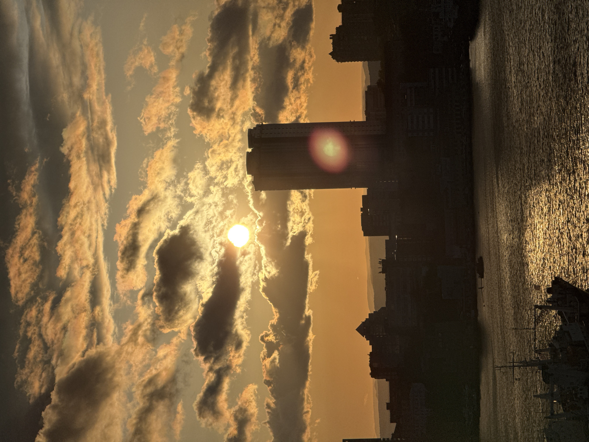 The image shows a sunset over a body of water, likely the sea. In the foreground, there are buildings in silhouette, indicative of a cityscape. The sun is visible through clouds, creating a bright glow. The clouds are illuminated with an orange-yellow hue. Part of a ship and its mast are visible at the bottom of the frame. The image location is Vladivostok, Russia.