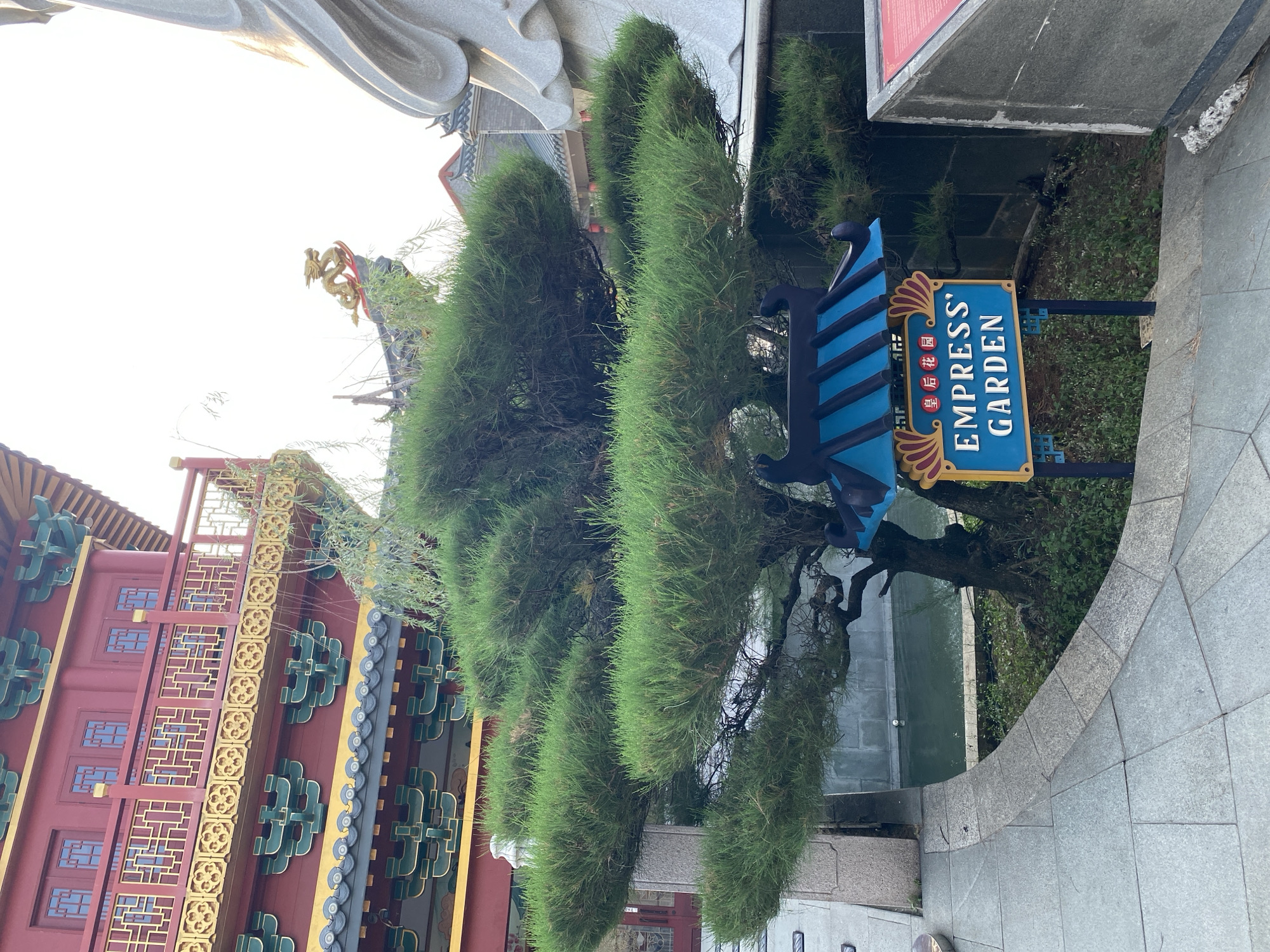 The image depicts a sign in front of a garden, possibly indicating an entrance or a landmark within a larger complex. The sign is blue with gold trim and reads "EMPRESS' GARDEN" in English, with Chinese characters above it. It has a decorative roof structure reminiscent of traditional East Asian architecture. Behind the sign, a lush green pine tree with sculpted branches is prominently featured. In the background, a multi-story red building with ornate gold and green accents and tiled roofs suggests a traditional East Asian architectural style. A large, light-colored statue, possibly a sculpture of a person, is partially visible on the right side of the frame. The ground is paved with light gray stone tiles. A body of water, likely a pond or canal, is visible behind the tree. The scene appears to be set in an outdoor environment, possibly a tourist attraction or a cultural park, given the signage and architecture. This location is within the "Dragonfly Village" complex at Pantai Indah Kapuk (PIK) in North Jakarta, Indonesia, which is known for its themed attractions and East Asian architectural influences.