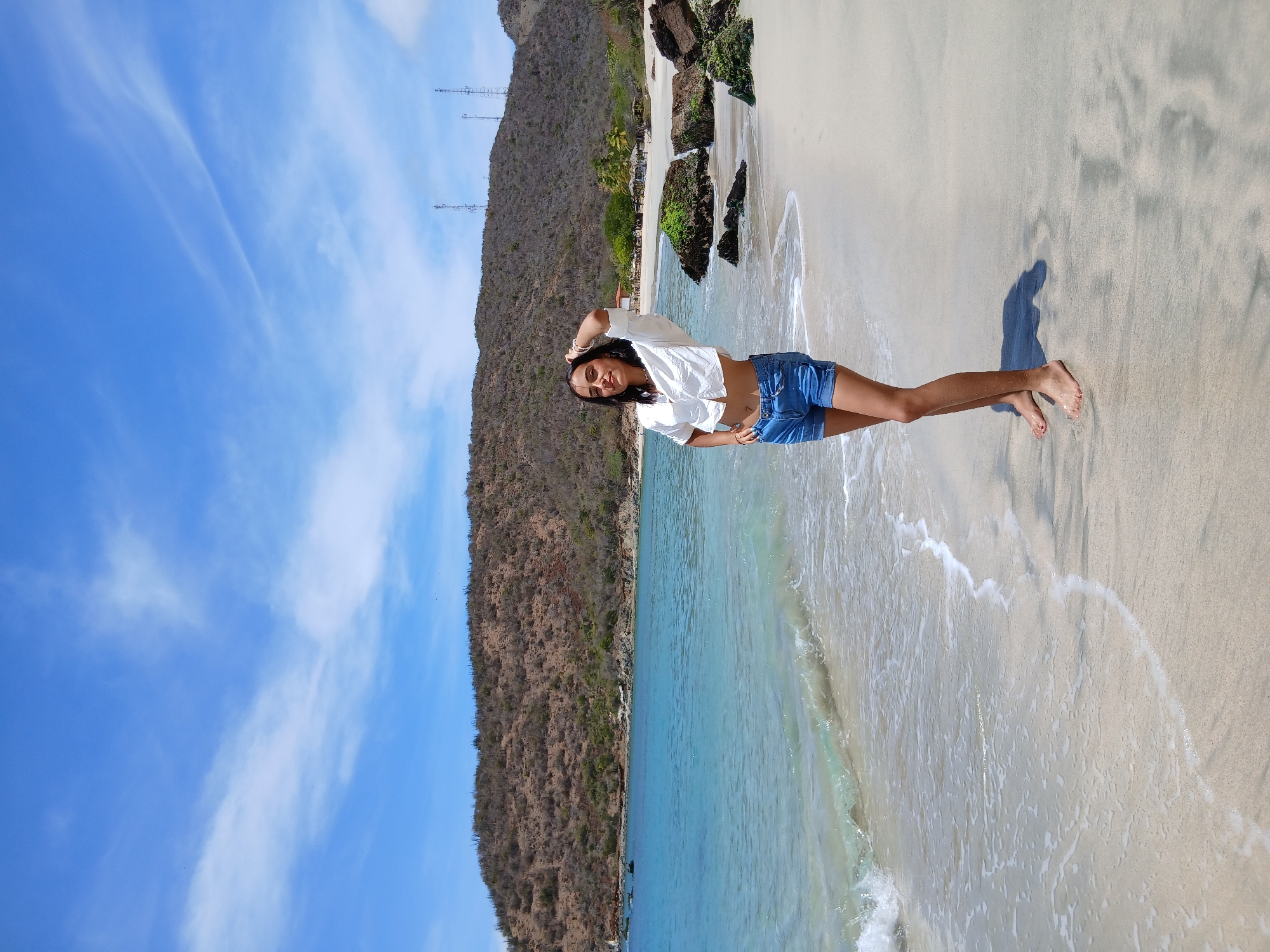 This image captures a vibrant beach scene in Juan Griego, Venezuela, on a bright, sunny day. The primary subject is a woman standing on the wet sand at the ocean's edge. She has long, dark hair and is dressed in a white, short-sleeved crop top knotted at the waist, exposing her midriff, and light blue denim shorts. Her bare feet, with red-painted toenails, are visible in the sand. She is smiling gently at the camera, posing with one arm raised and her hand near her head, and the other hand resting on her hip. Her shadow is cast on the sand behind her, indicating a high sun position.
The immediate foreground features light-colored sand with patterns left by receding waves, and the clear, turquoise water of the ocean. Gentle white-capped waves are seen breaking and pulling back from the shore. Beyond the water, a rugged coastline rises, characterized by a large hill or mountain covered in dry, sparse, brown vegetation. Several tall communication towers are visible atop this distant hill, silhouetted against the bright blue sky. The sky itself is expansive and clear, with only a few thin, wispy white clouds. Closer to the water's edge on the coastline, some rocks are visible, partially covered in green algae or moss. The overall impression is one of a cheerful and relaxed tropical beach environment. There is no visible text in the image.