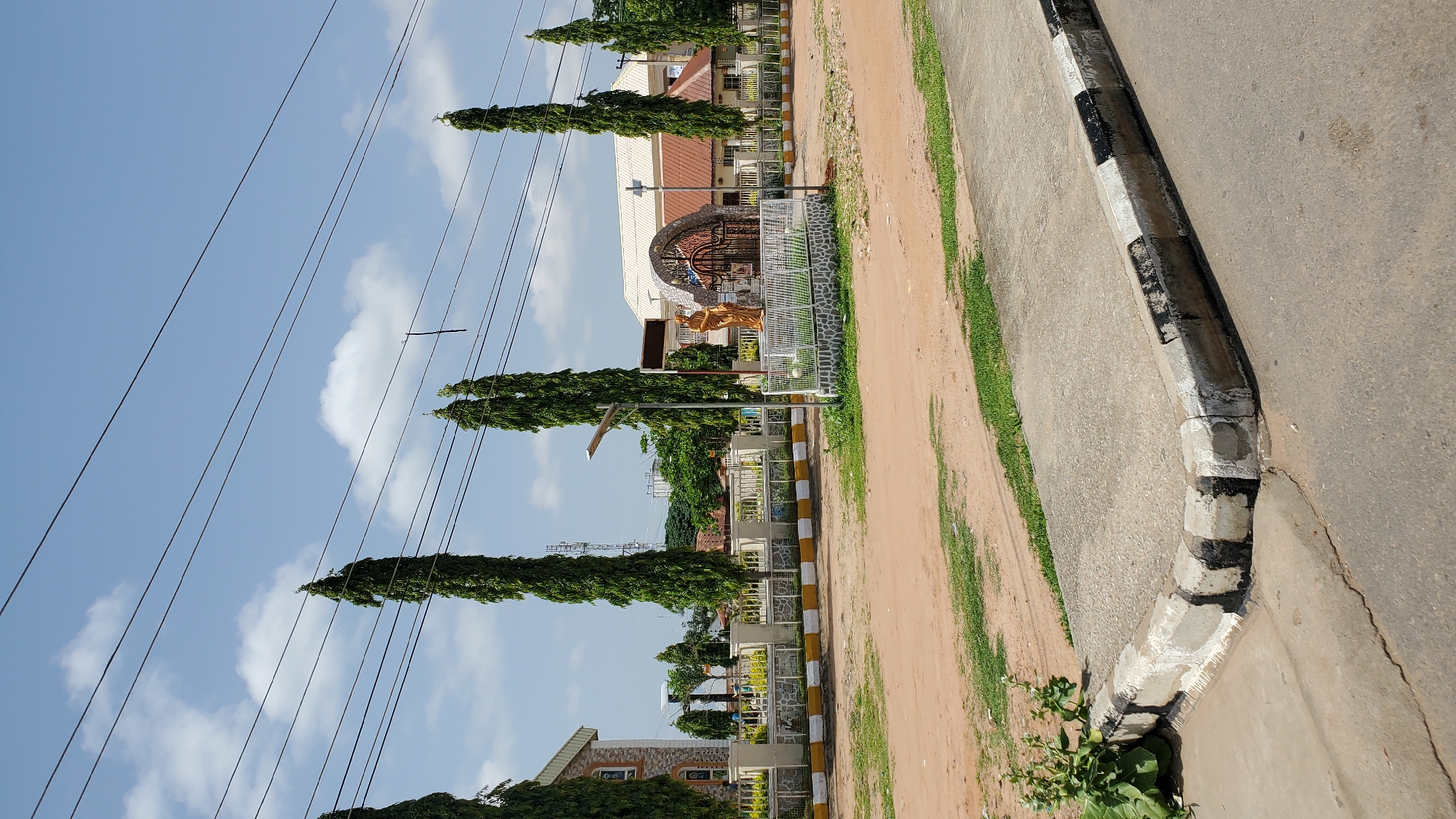 In the city of Jalingo, Nigeria, this image captures a street view facing a white and yellow fenced perimeter with tall, slender trees lining the interior. Beyond the fence, a building with a light-colored facade and brown roof is visible on the left, while a larger structure with a white roof and brown accents is situated towards the right. A decorative arched gateway with intricate metalwork stands at the center of the fenced area, flanked by a golden statue of a person on the left and a white structure on the right. Two lampposts are positioned within the compound. The foreground shows a dirt road with patches of grass and a concrete curb painted with alternating black and white stripes. Overhead, several power lines stretch across a bright blue sky dotted with white clouds. The scene depicts a public or religious space within an urban environment.