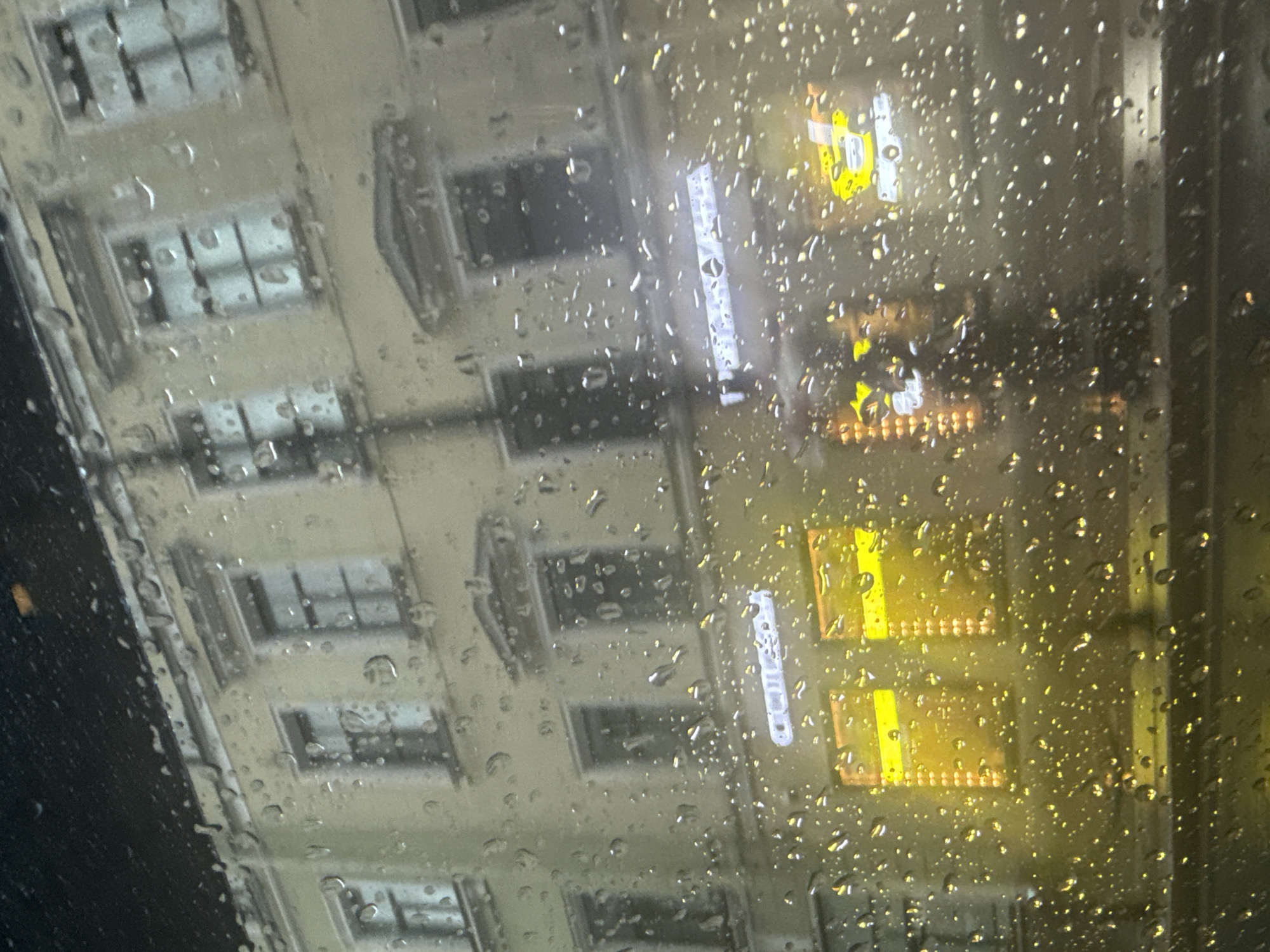 The image captures a street scene at night in Saint Petersburg, Russia, viewed through a window covered in raindrops. A multi-story building with a pale facade dominates the background, featuring rows of windows on multiple levels. The windows are mostly dark, but some on the lower floors are illuminated with bright yellow light, suggesting shops or businesses within. A neon sign, partially obscured by raindrops, is visible on the building's lower level, with some letters appearing to be in Russian. The overall impression is one of a wet, urban environment after dark, with the raindrops distorting the view of the building and the street.