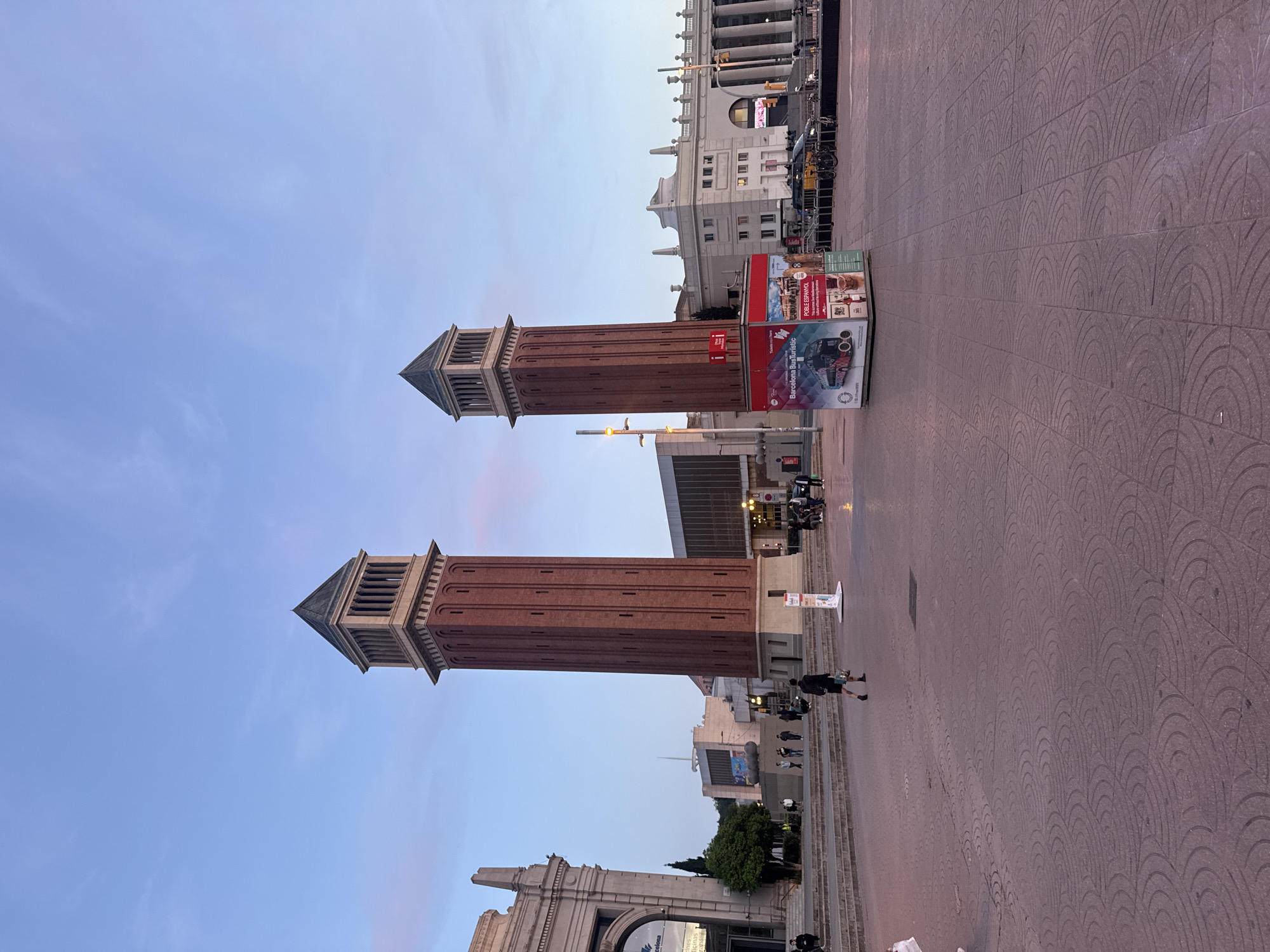 The image shows the Venetian Towers in Barcelona, Spain, taken from a slightly elevated position. Two red brick towers with a gray crown-like top structure are prominently displayed against a light blue sky. The towers flank a wide pedestrian thoroughfare, paved with a patterned surface of semi-circular designs. A few people are seen walking on the thoroughfare. Several buildings are visible in the background, including one with a colonnaded facade and another with a modern design. Several advertisement stands can be seen on the pavement.
