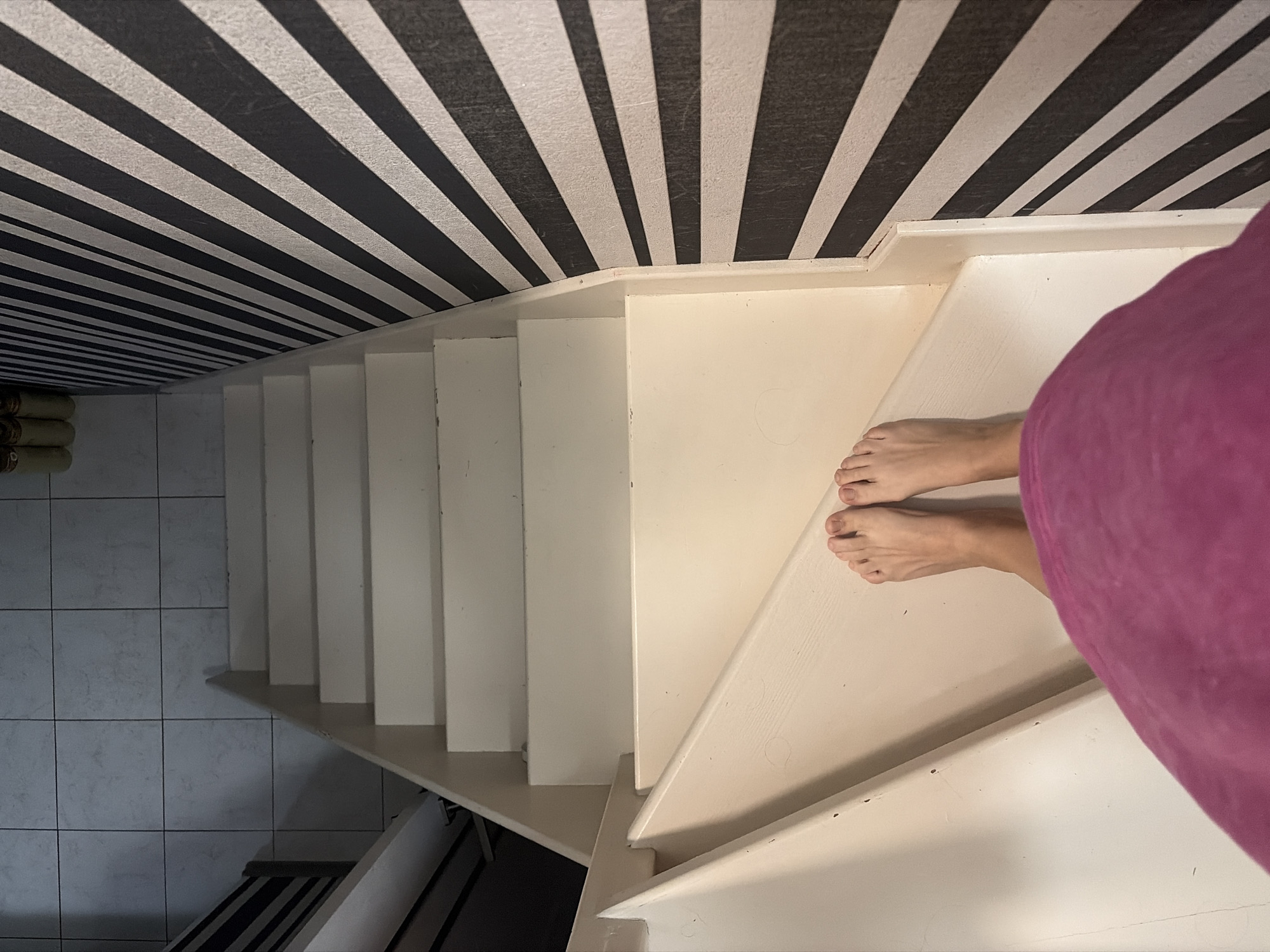 The image shows a top-down view of a person's feet standing on a carpeted staircase. The staircase is cream-colored and leads downwards. To the right of the staircase, there is a wall covered in black and white striped wallpaper. The floor at the top of the stairs appears to be tiled. The person is wearing a pink garment that covers their lower body and legs. The image does not contain any visual information that would indicate the location is in Sochi, Russia.