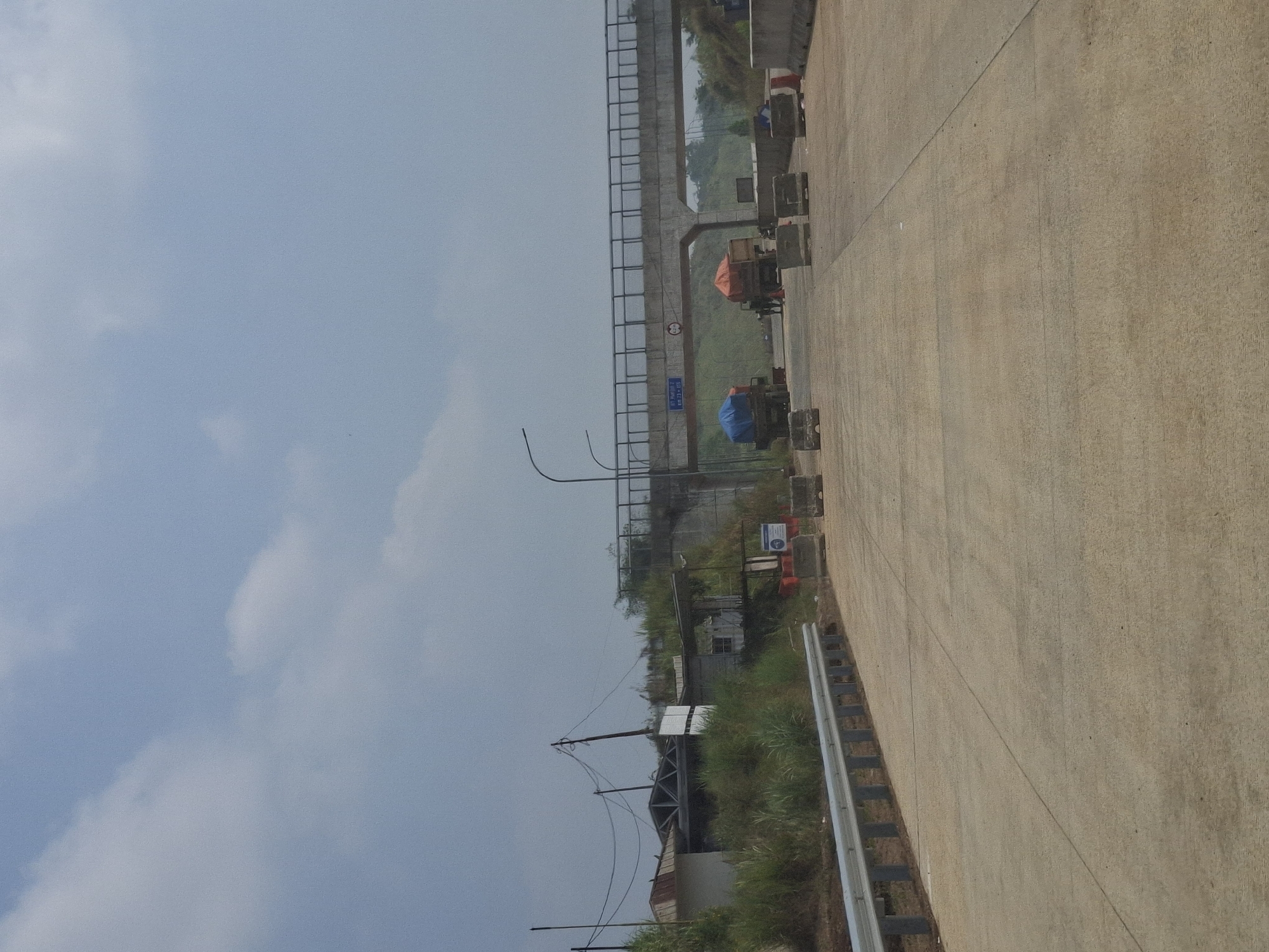 The image depicts a highway scene in Parungkuda, Indonesia, under a partly cloudy sky. In the foreground, a concrete highway stretches into the distance, bordered by a metal guardrail on the left. Several concrete barriers are strategically placed on the highway. In the middle ground, a pedestrian bridge spans the highway. Two trucks, one with a blue tarp and another with an orange tarp, are visible on the highway beneath the bridge. On the left side of the highway, beyond the guardrail, there are some low-rise structures, vegetation, and utility poles with overhead wires. A sign with blue text is attached to the pedestrian bridge. The environment appears to be semi-urban or industrial.