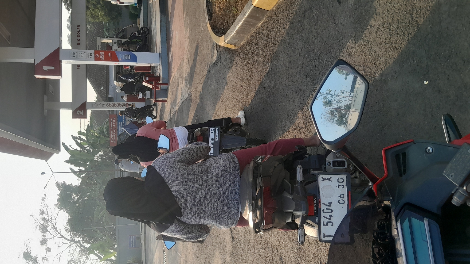 This image depicts a gas station in Subang, Indonesia. Two women on motorcycles are waiting in line. The woman in the foreground is wearing a grey knitted sweater and red pants, with a black hijab. The woman behind her is wearing a pink shirt and a black hijab. The gas station has pumps labeled with numbers 1 and 2, and signage indicating "BIO SOLAR". In the background, other people on motorcycles and a white van are visible. The environment appears to be an outdoor setting with trees and a clear sky. The license plates of the motorcycles are visible, with one reading "T 6909 ZG" and another "T 5404 X C6 3G". The scene suggests a typical day at a gas station in Indonesia, with a queue of vehicles waiting to refuel.