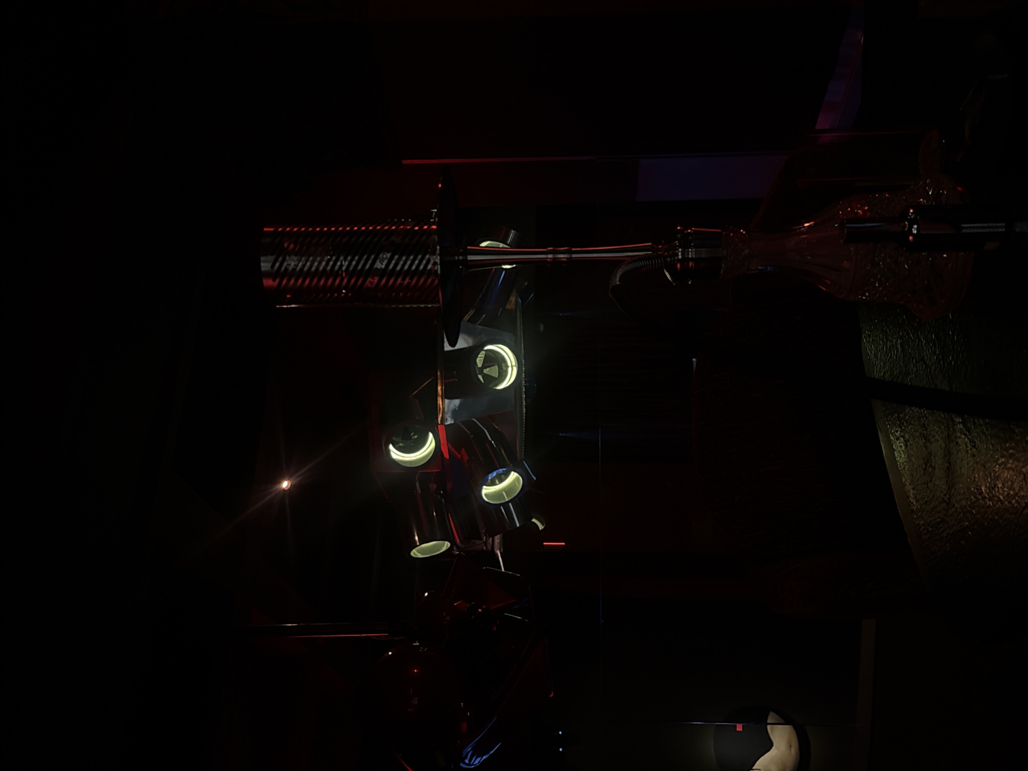 The image depicts a dimly lit interior, possibly a nightclub or lounge, with a strong focus on atmospheric lighting and decorative elements. In the center of the frame, a complex lighting fixture hangs, featuring multiple circular lamps with bright, yellow-white illumination. The fixture itself appears metallic and angular. To the right of this lighting unit, a tall, cylindrical object with a textured, ribbed surface, possibly a speaker or decorative column, is visible, accented by red light. In the lower right quadrant, a detailed hookah is prominent, with its glass base filled with a reddish liquid, and its metallic stem extending upwards. The environment is dark, with hints of red and blue lighting reflecting off surfaces. A circular object with a light beige surface and a red accent is partially visible in the lower left. The overall impression is one of a stylized, modern entertainment venue, likely in Moscow, Russia, given the context of the prompt.