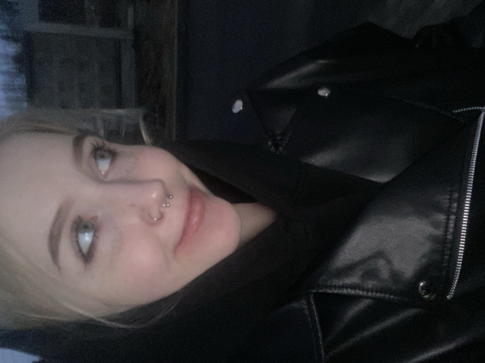 A close-up, slightly angled shot shows a fair-skinned individual with light blonde hair, possibly a young woman, wearing a black leather jacket over what appears to be a black hooded garment. The person has a double nose piercing on one side of their nose. The background is dark and indistinct, but a structure reminiscent of a building or wall is vaguely discernible. It's not possible to confirm the location as Izhevsk, Russia, based on visual information alone.