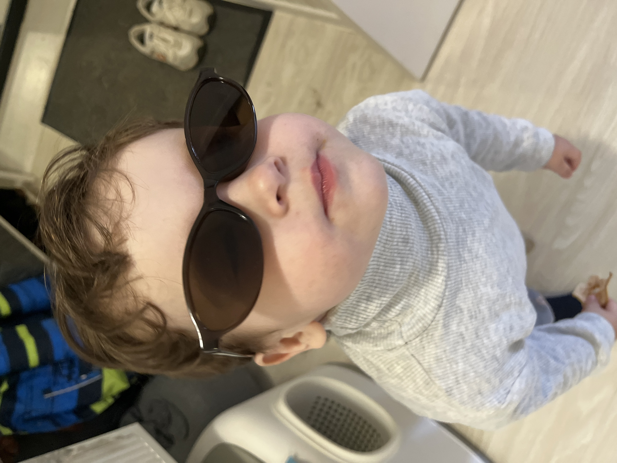 A child wearing sunglasses and a gray turtleneck stands in a room with light-colored floors. The background includes a mat with two pairs of shoes, and a blue and yellow jacket. The child is holding something small in their right hand. A white object is near the child's torso. There are no indicators that the image was taken in Chaikovsky, Russia.