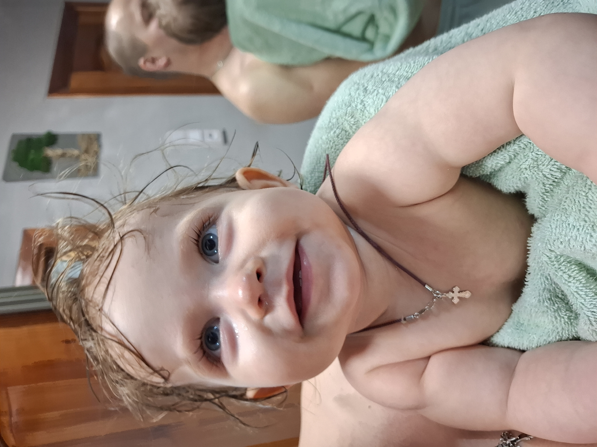 A close-up shot captures a baby wrapped in a light green towel, possibly after a bath. The baby has wet, light brown hair and blue eyes, and wears a necklace with a cross pendant. Visible in the blurred background is a person with a beard, also wrapped in a towel. The location is identified as Chaikovsky, Russia. A wooden structure is partially visible on the lower-left and top-left of the frame. Artwork is seen hanging on the wall.