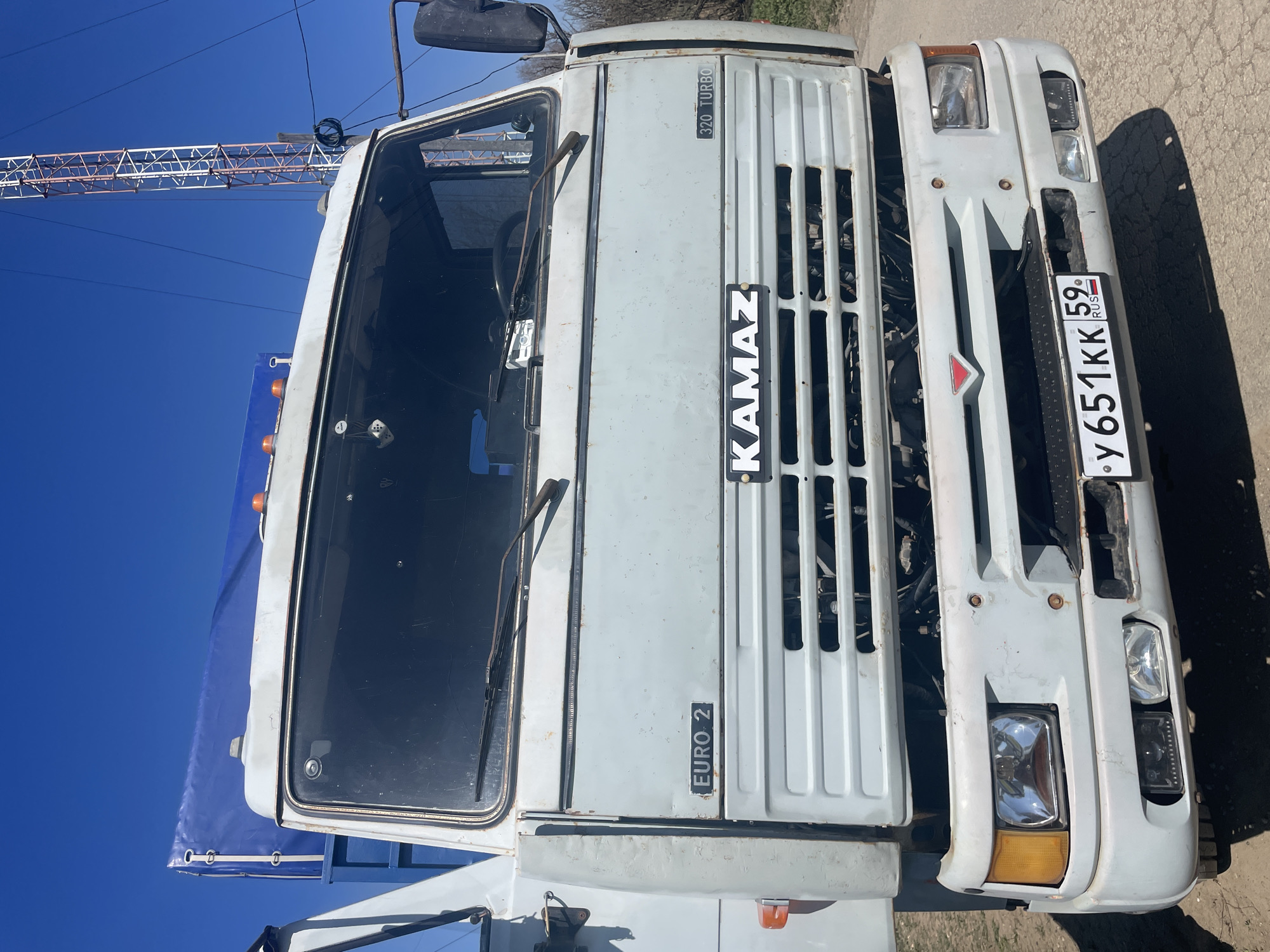 The image shows the front of a white KAMAZ truck. The license plate reads "y 651 KK 59 RUS," indicating it's registered in Russia, specifically the Perm Krai region (code 59). The truck is labeled "320 Turbo" and "Euro 2," suggesting it's a model from the late 1990s or early 2000s. The location appears to be in the city of Поедуги, Russia. The background includes a blue sky and a metal structure, possibly a crane or power line support. The truck's condition suggests it's an older vehicle, with visible wear and tear.