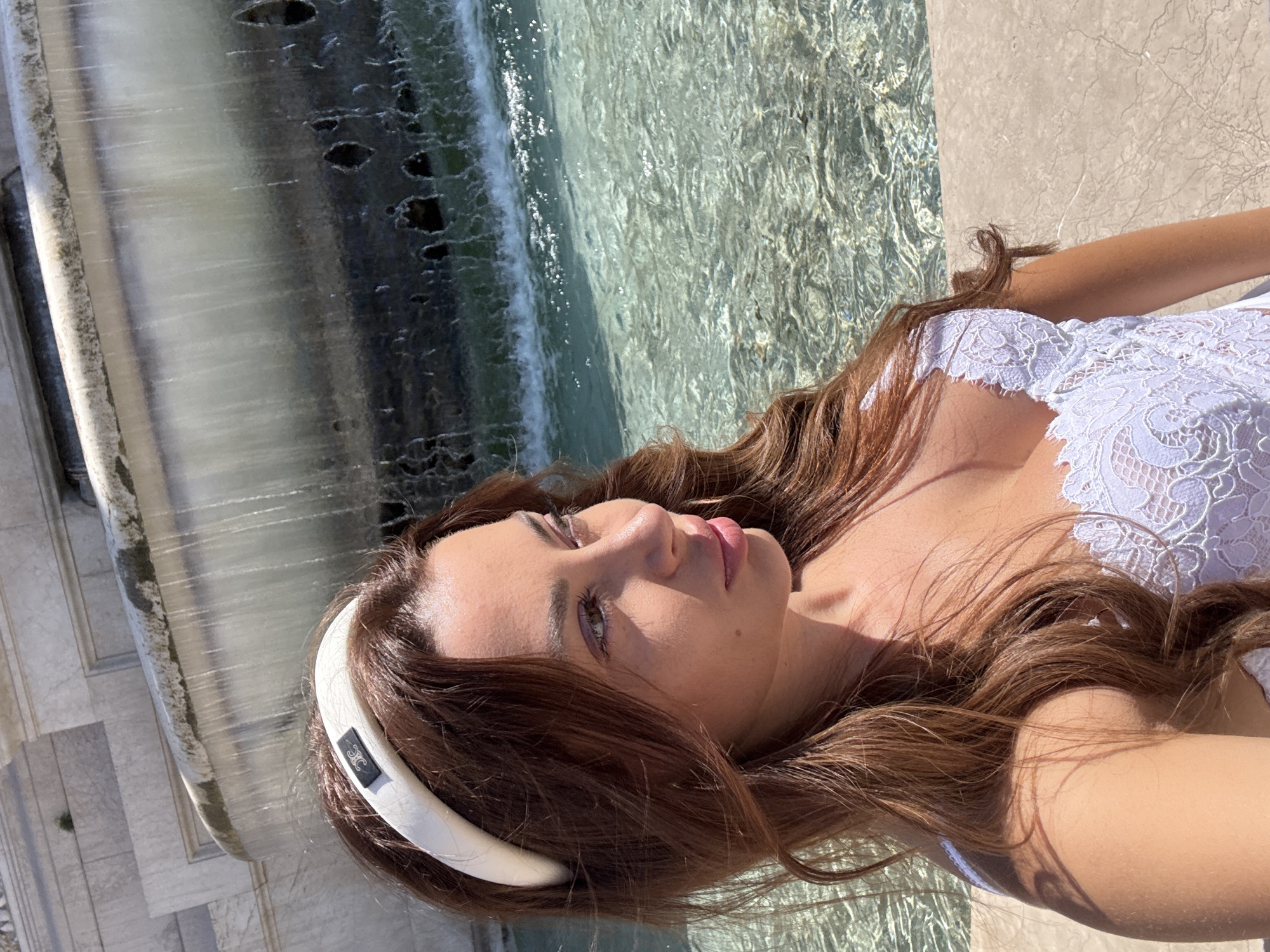 A medium shot shows a young woman with long, wavy brown hair and a white lace top. She is wearing a white padded headband with a black logo. She is positioned in the foreground, with a fountain behind her. The fountain features water cascading over a tiered stone structure. The water is clear and reflects the sunlight, creating a shimmering effect. The woman is looking to her right, with her profile visible. The scene appears to be outdoors, bathed in sunlight. The stone structure in the background resembles the Trevi Fountain in Rome, Italy, known for its Baroque architecture and abundance of water features.