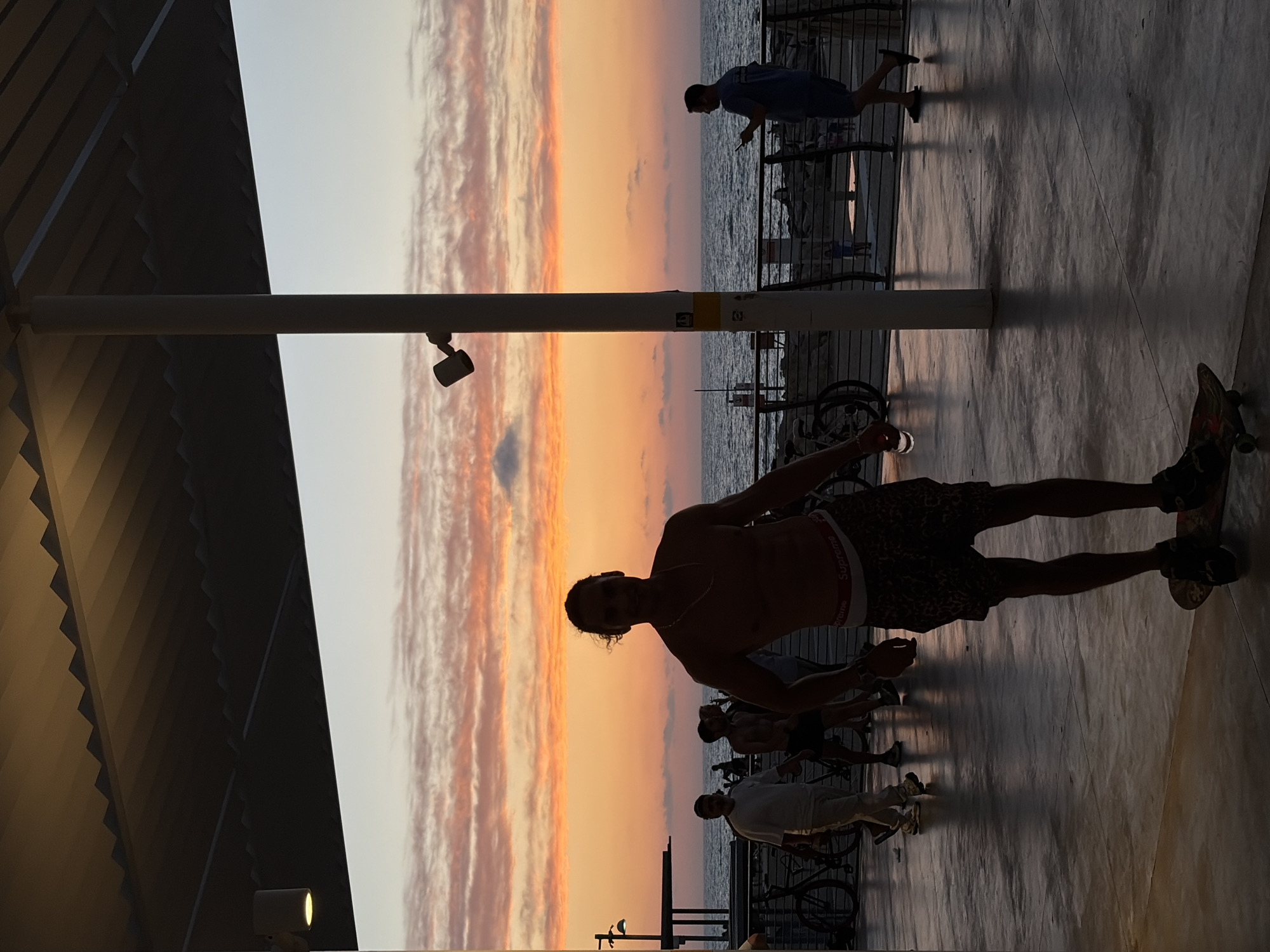 A wide-angle, slightly off-center shot shows a group of people silhouetted against a sunset in what appears to be Tel Aviv, Israel. The sky is awash in hues of orange, pink, and white, reflecting off the sea visible in the background. A metal railing runs horizontally across the center of the image, separating the foreground activity from the ocean.
In the foreground, a man with curly hair is standing on a skateboard, shirtless and wearing patterned shorts. His silhouette is prominent, suggesting he is closer to the camera. Several other people are present, either standing or walking, near bicycles.
The scene is partially under a modern structure with a slanted, slatted roof, casting shadows on the ground. A vertical pole with a light fixture is positioned to the left, further contributing to the interplay of light and shadow. The ground is a paved surface, reflecting the ambient light.