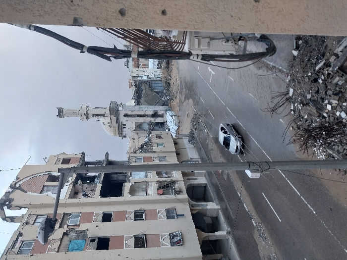 This image, viewed from an elevated vantage point, depicts a scene of extensive urban destruction in Khan Yunis, Palestinian Territory, under an overcast sky.
The central focus is a street flanked by heavily damaged multi-story buildings and piles of rubble. On the left, a large building shows severe structural compromise, with gaping holes, shattered windows, and large sections of its facade missing, revealing exposed interiors. Further down the street, a mosque with a prominent minaret and dome is visible, also appearing to have sustained damage but largely standing amidst the devastation.
The paved road in the foreground and middle ground has a white sedan driving on it, or possibly parked, suggesting some limited activity. However, the street sides are littered with significant piles of concrete debris, twisted rebar, and other wreckage, particularly on the right side of the frame. Overhead wires and a utility pole are visible, running across the scene. The immediate foreground shows a concrete surface, possibly a balcony or roof edge, with what appear to be impact marks or bullet holes.
The scene conveys the aftermath of severe conflict or a major destructive event, showcasing widespread damage to an urban environment. The time of day appears to be daytime, under a dull, cloudy sky. No discernible text is visible in the image.
