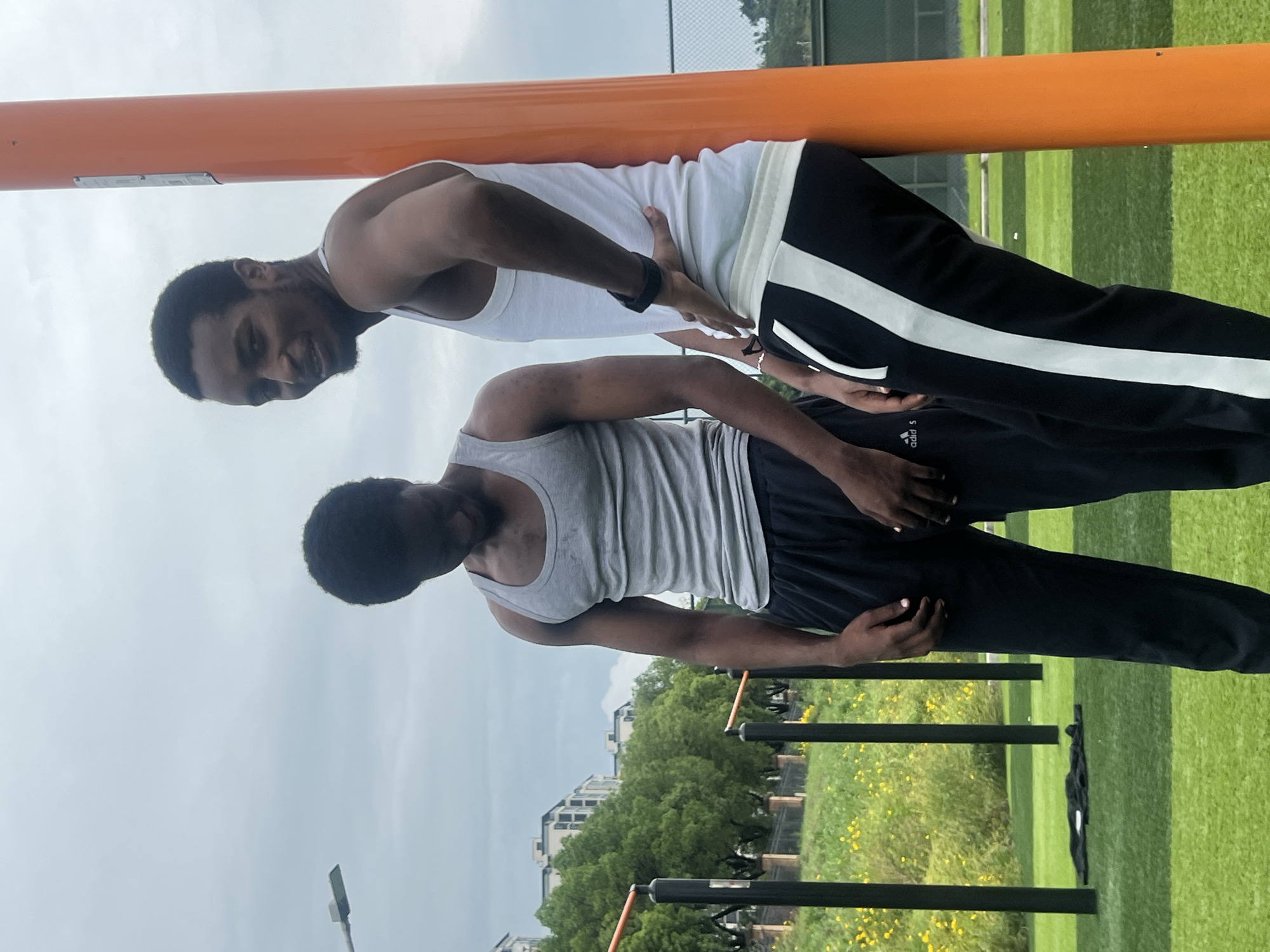 Two African American males are outdoors. The top man is wearing a white tank top and black Adidas track pants with white stripes. The bottom man is wearing a grey tank top and black Adidas track pants. Both men are holding onto a horizontal orange bar. The background includes green grass, yellow flowers, black metal bars, trees, and a tall building. A street lamp is visible in the background. The location is Jiangjin, China.