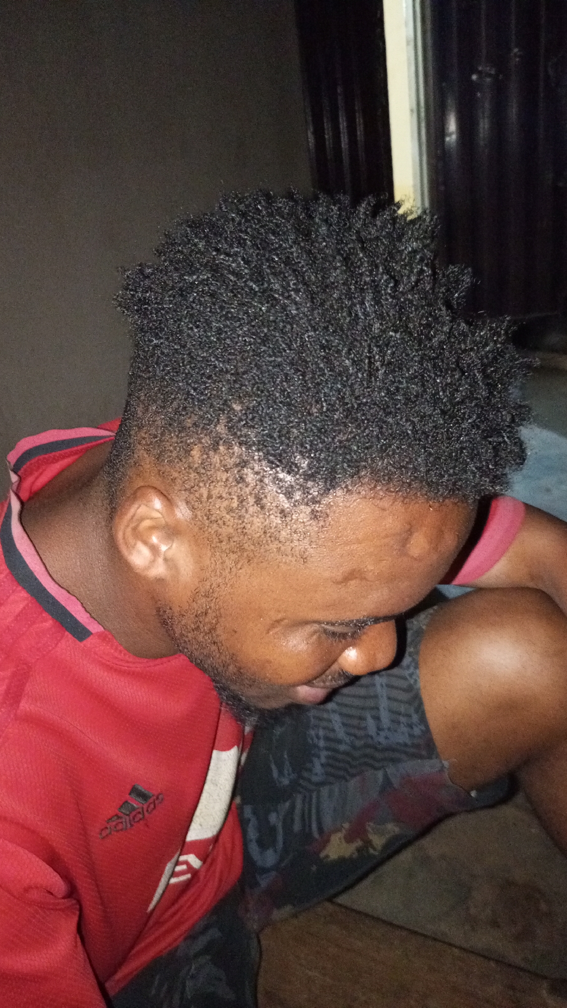 A close-up, indoor shot shows a man in Sabon Tasha, Nigeria, looking down. He wears a red Adidas shirt with black stripes and a white logo. His hair is short, dark, and styled with texture. He has a short beard. His shorts are patterned in gray and black. The background includes a partially visible doorframe and wall. The lighting is dim.