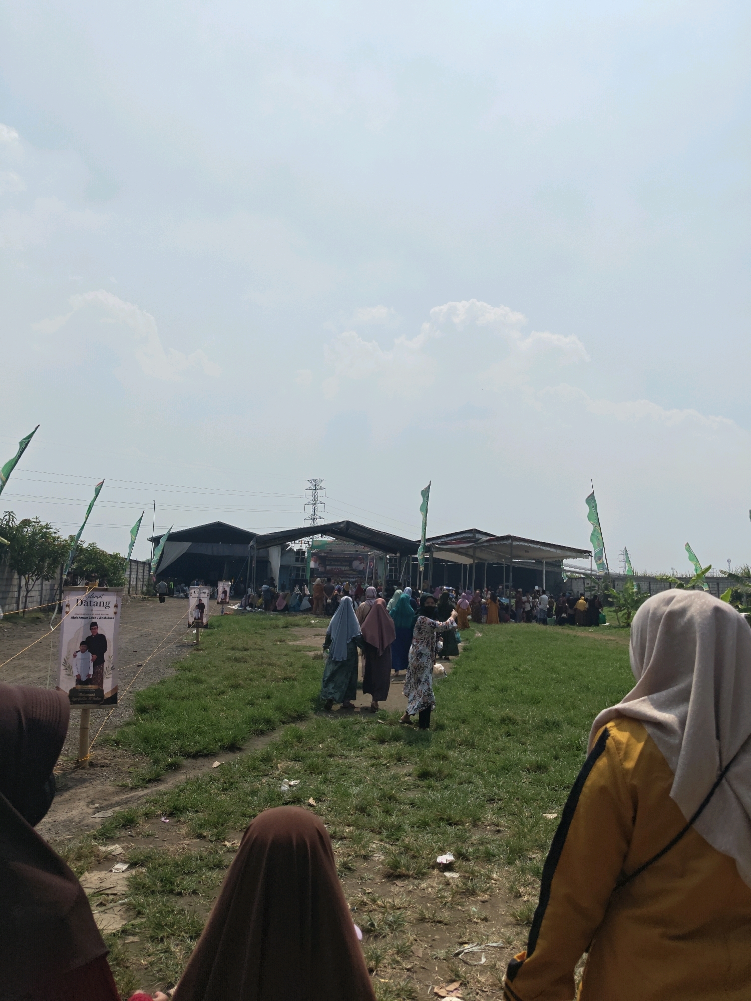 The image depicts an outdoor gathering in Bangsal, Indonesia. In the foreground, three individuals are partially visible, their backs to the camera, wearing headscarves. The central figure wears a yellow and black jacket, while the other two wear brown headscarves. A banner in the mid-left of the image reads "Selamat Datang" and features portraits of two men. Green flags are prominently displayed throughout the scene, attached to poles and fluttering in the wind. In the background, a crowd of people, many wearing headscarves, are gathered around several low-lying buildings with open sides. Some individuals are seated on the ground, while others appear to be standing or walking. The environment is characterized by dry, dusty ground and patches of green grass. A tall electrical tower is visible in the distance, against a pale blue sky with scattered white clouds. The overall impression is of a community event taking place in a semi-rural setting.