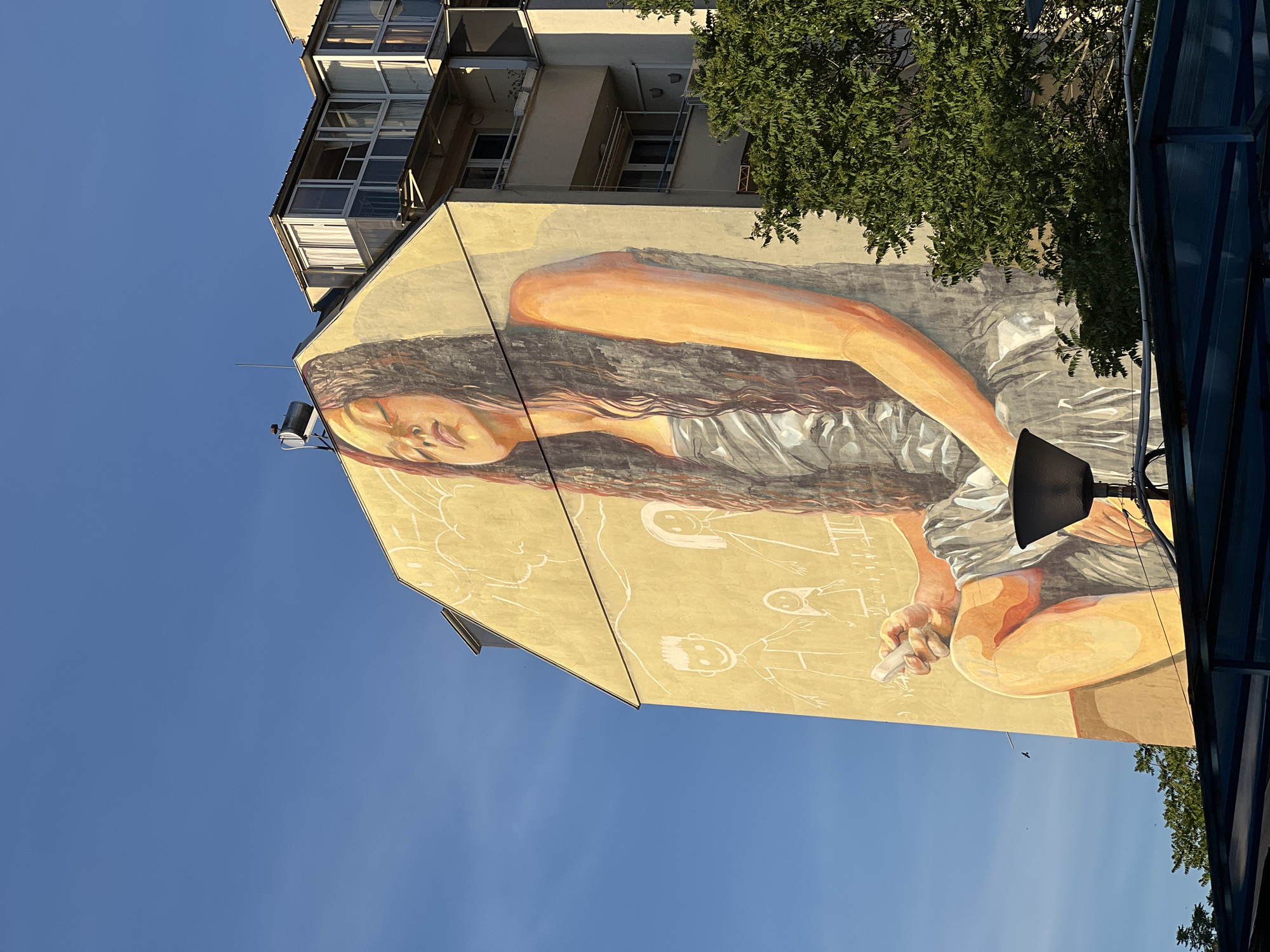 A mural in Sofia, Bulgaria, depicts a woman with long brown hair lying on her side, wearing a silver-grey dress. The mural covers the side of a tall, multi-story building. Below the woman are simple drawings of children. The background is a yellow-tan color. The sky is visible in the lower-left corner. A tree and part of a metal structure appear on the right side of the image.
