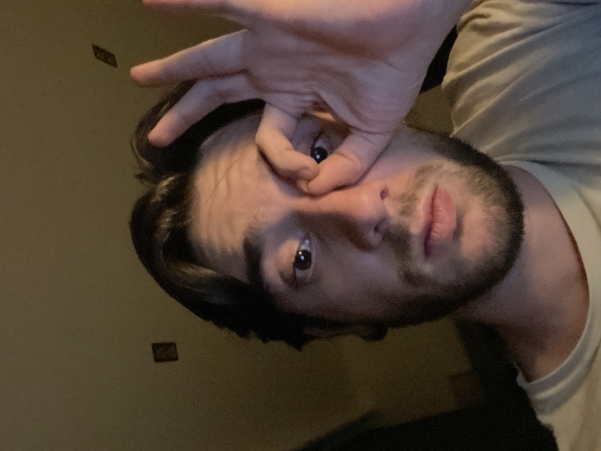 The image is a close-up selfie of a young man with dark hair and a beard, holding his right hand up to his face, with his index finger and thumb forming a circle around his right eye. He is wearing a light-colored t-shirt. The background is a dimly lit, plain wall with two small, dark square objects mounted on it. The lighting suggests an indoor environment, possibly at night. There is no identifiable information in the image to place it in Huixquilucan de Degollado, Mexico.