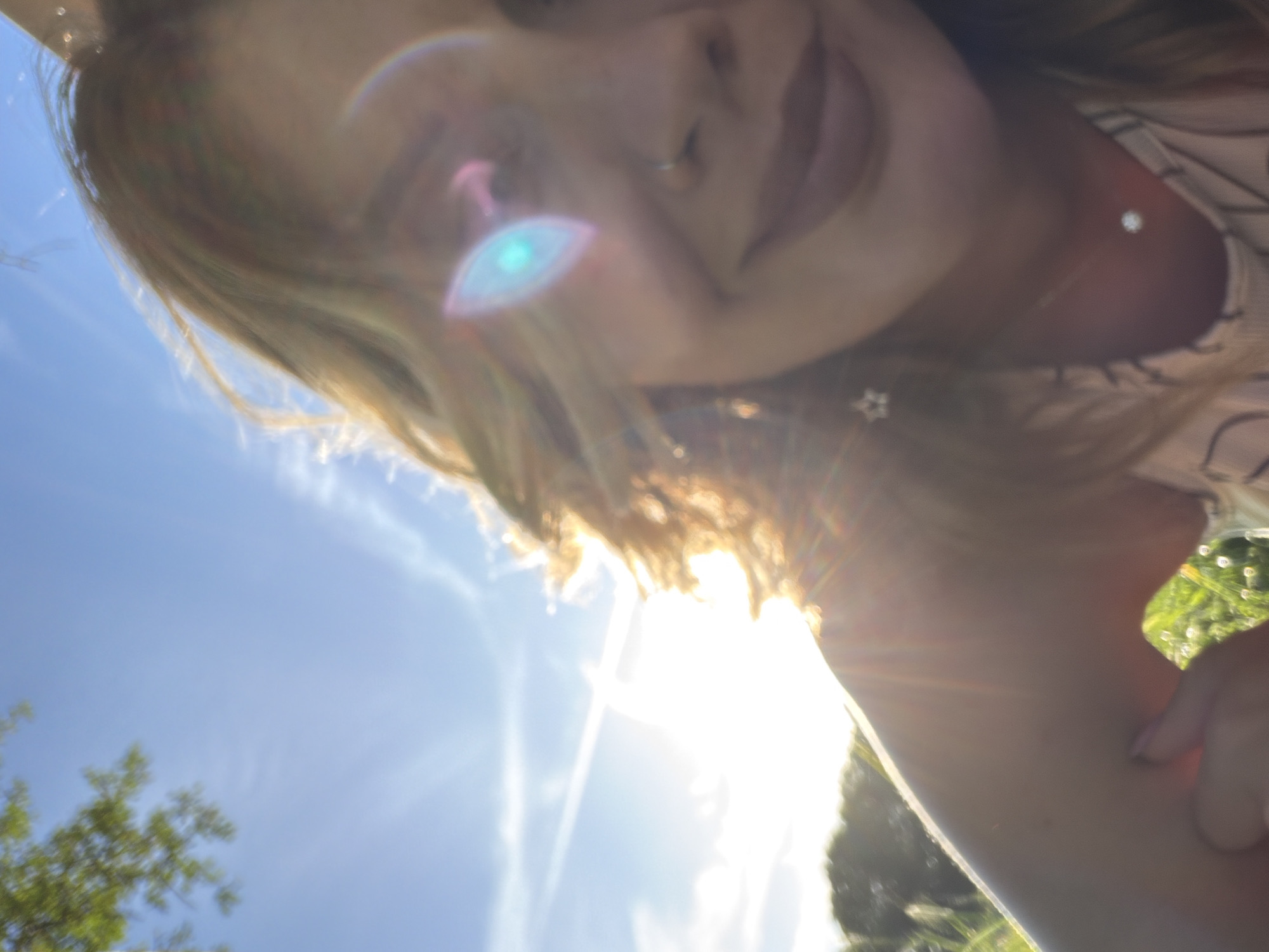 The image shows a close-up selfie of a person with fair skin and light brown hair. The person has a nose ring and is smiling slightly. The background includes a bright blue sky with some tree branches visible in the lower left corner. A significant amount of sunlight is present, causing lens flare and obscuring details in the background. The location cannot be determined from this image.