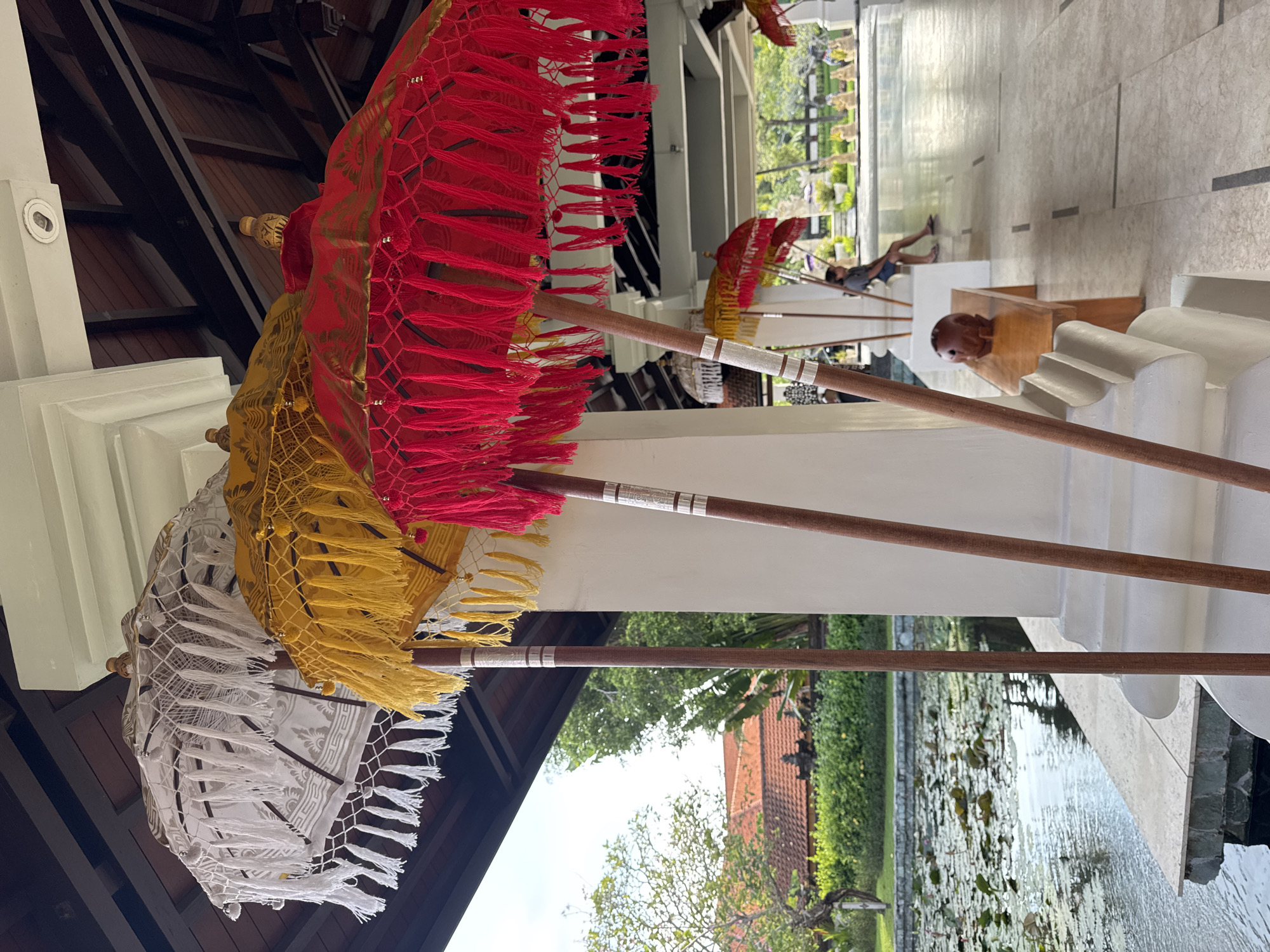 A variety of colorful decorative umbrellas, primarily red, yellow, and white, are stacked near a walkway. The umbrellas are adorned with fringe and have wooden poles. The walkway is adjacent to a pond with lily pads, suggesting an outdoor or courtyard setting. In the background, a person is seen lounging in a seated position, near what appears to be statues. The image captures a moment in a tropical setting, likely in Kuta Selatan, Indonesia, as per the prompt.