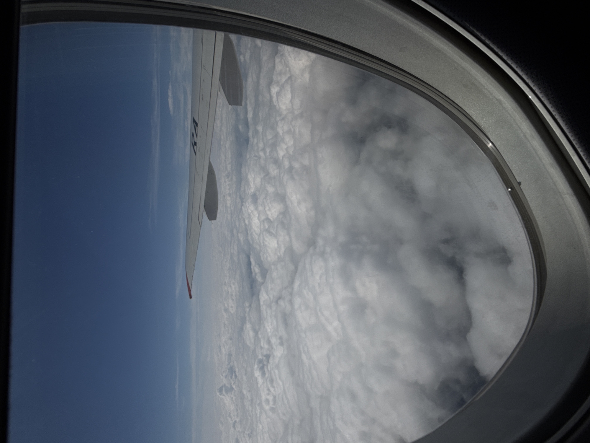 The image, taken from an airplane window, displays a vast expanse of white clouds stretching to the horizon, bathed in sunlight. The sky above is a deep, clear blue. A portion of the airplane's wing is visible in the upper right quadrant, with the letters "RA" printed on its surface. The view is framed by the dark interior of the airplane cabin. The location in Staroe Mart'yanovo, Russia is not discernible from the image.