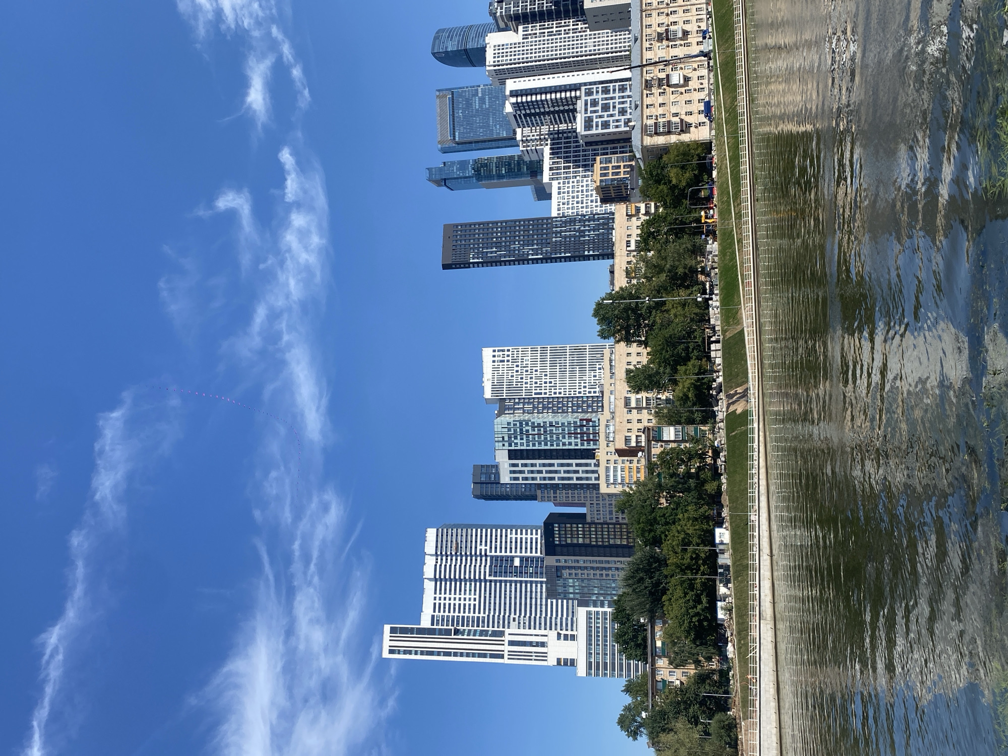 This is a wide, horizontal shot of a cityscape in Moscow, Russia, likely during the day. In the foreground, a calm river reflects the sky and the buildings. A white railing runs along the embankment. Behind the embankment, a line of trees and then a row of buildings are visible. Dominating the background is a modern skyline of numerous skyscrapers of varying heights and architectural styles, with some buildings featuring reflective glass facades. The sky is a clear, bright blue with scattered, wispy white clouds. A thin line of small, pink, kite-like objects is visible in the upper right portion of the sky, forming a curved arc. The scene appears to be peaceful and urban.