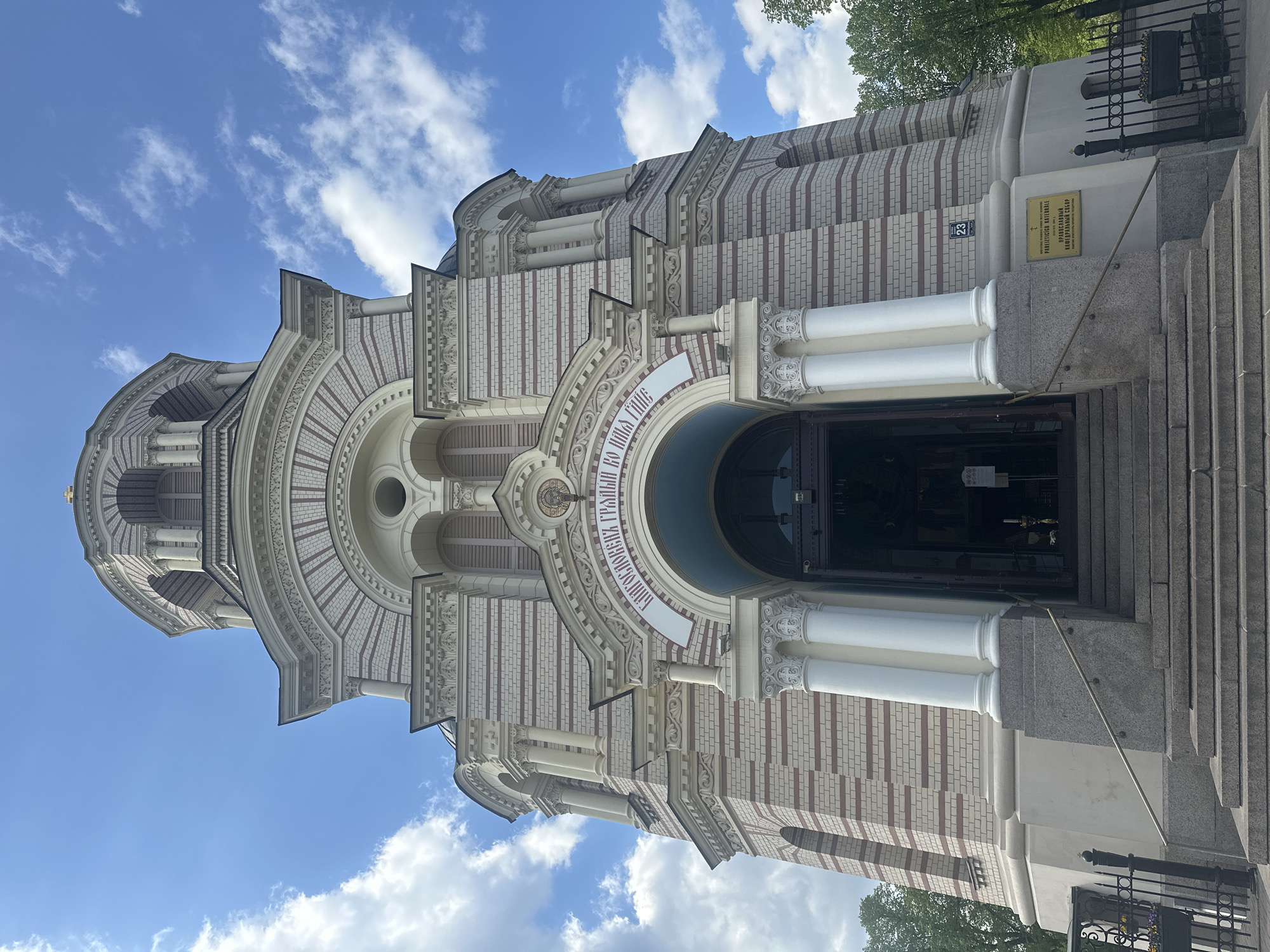 The image showcases the Nativity of Christ Orthodox Cathedral in Riga, Latvia. The building's facade, characterized by a light-colored brick design with alternating red brick accents, dominates the view. Ornate archways and decorative columns add to its architectural complexity. A central entrance, framed by white columns and a semicircular arch, invites entry. Above the archway, Cyrillic script adds a cultural element to the scene. A plaque with Cyrillic text is visible on the side, and a number "23" is attached to the building. The sky is partially visible, featuring a blue backdrop with scattered white clouds. A fence, the street, and trees are at the bottom of the picture. The image is taken at an angle, looking upwards at the cathedral.