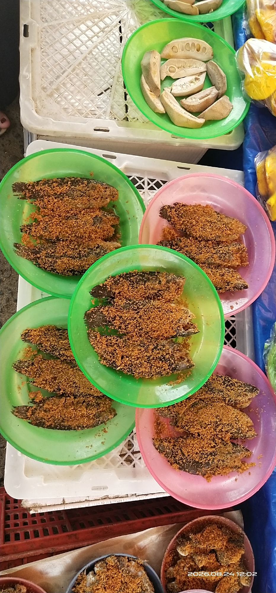 In Tamiang Layang, Indonesia, a market stall displays several small, fried fish seasoned with a granular coating, arranged on colorful plastic plates. The plates, green and pink, are stacked on white plastic crates. Some of the fish are visible in the foreground, covered in the seasoning. In the upper right portion of the image, a green bowl contains slices of a dried fruit, possibly jackfruit. The background is a blur of market activity, with hints of other produce and market structures. The overall scene suggests a bustling local food market environment.