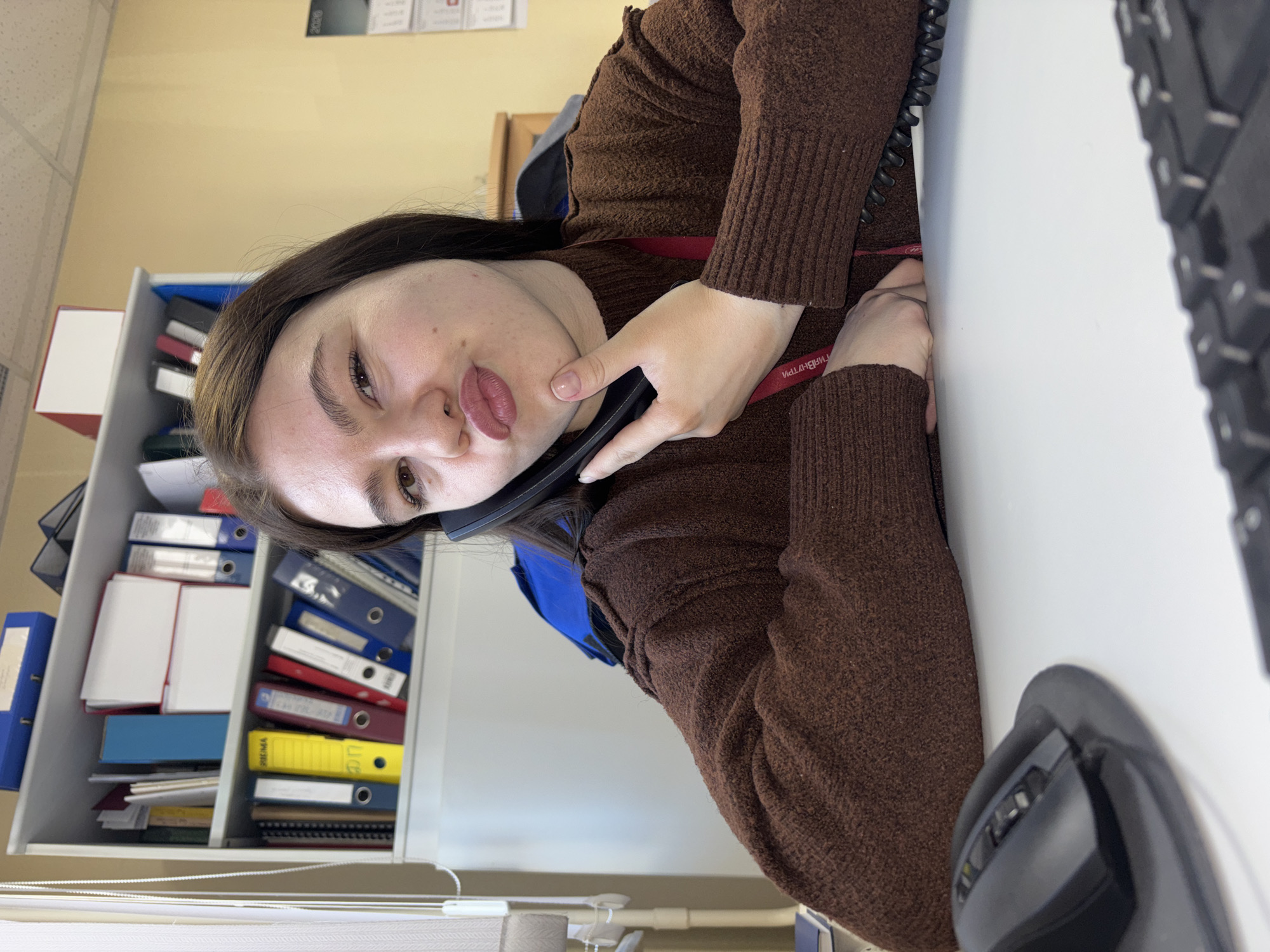 A young woman in a brown sweater is holding a telephone receiver to her ear while seated at a desk. The desk contains a keyboard and a computer mouse. To her left is a shelf filled with binders and documents. The woman is wearing a red lanyard. The photograph appears to be taken in an office environment. Based on the lanyard inscription in Russian, the location is likely Perm, Russia.