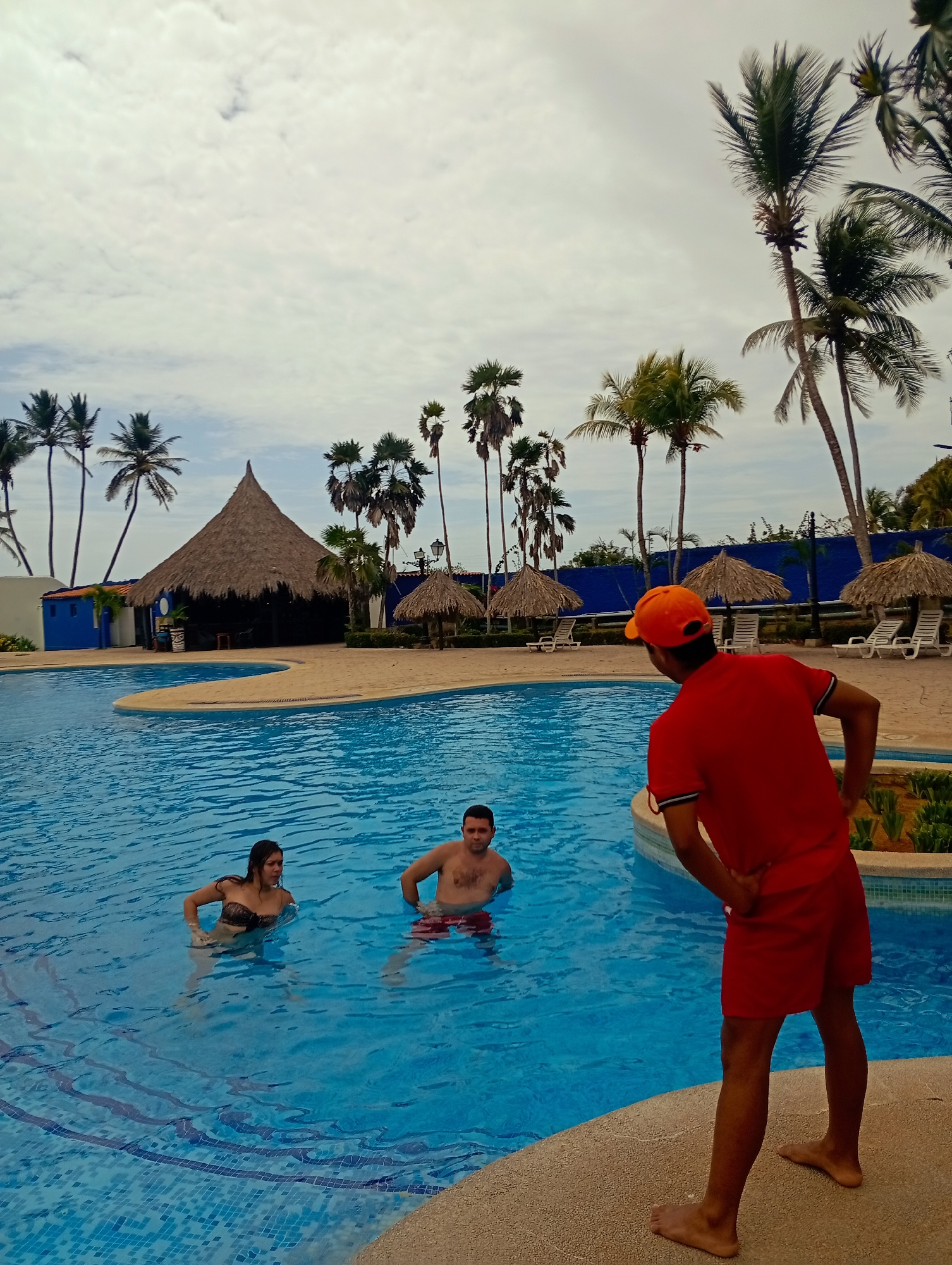 This image captures a vibrant, yet overcast, daytime scene at a tropical resort pool, specifically identified as Playa El Agua, Venezuela.
In the foreground, a man, seen from behind, stands barefoot on the pool deck. He wears a bright red t-shirt, red shorts, and an orange baseball cap, with his hands placed on his hips, suggesting a relaxed or supervisory stance. He faces two individuals in the blue-tiled swimming pool.
Within the irregularly shaped pool, a woman on the left is submerged up to her chest, wearing a dark patterned bikini top. To her right, a man is also chest-deep in the water, wearing red swim trunks. Both appear to be attentive to the man on the deck, suggesting an ongoing conversation or interaction. The pool water is clear and blue, revealing the mosaic tiling on the bottom.
The midground features the edge of the pool with a light brown, textured deck. In the background, a large, traditional conical thatched-roof structure, known as a churuata or palapa, stands prominently, likely serving as a bar or communal area. To the right of this main structure, several smaller thatched parasols (palapas) are visible, providing shade over white lounge chairs arranged on the deck. Numerous tall palm trees, characteristic of a tropical environment, are scattered throughout the scene, framing the structures and pool area. A low blue wall or fence with a black railing runs along the farthest background.
The sky is heavily overcast with light grey clouds, indicating a cloudy day, though still bright. No discernible text is visible in the image. The overall mood appears calm and leisurely, typical of a resort setting.