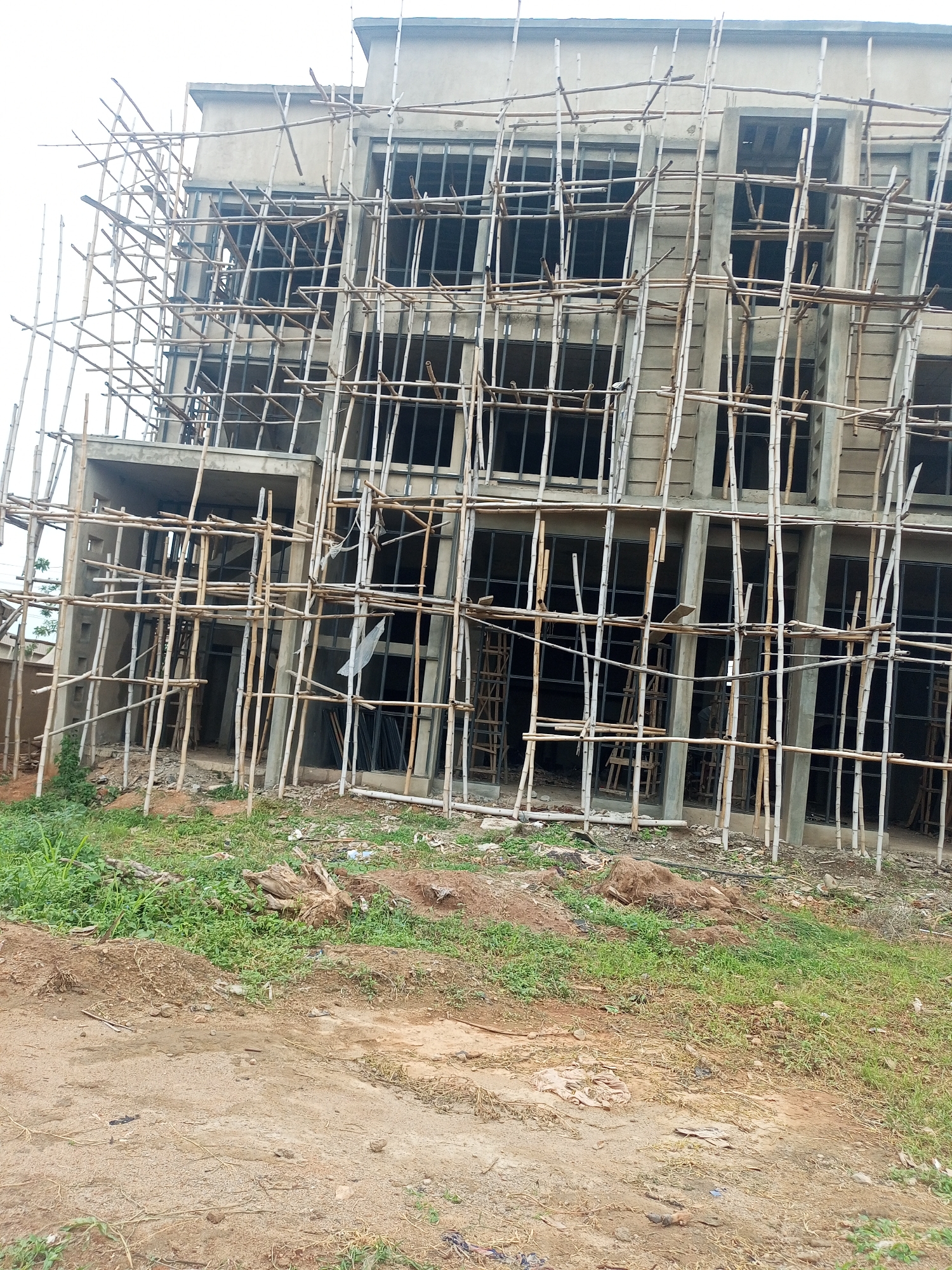 The image shows a partially constructed multi-story building in Oshogbo, Nigeria. The building's exterior is gray and appears to be made of concrete. Window frames are installed, but no glass is visible. Bamboo scaffolding surrounds the building, providing access for construction workers. The ground is a mix of dirt, sand, and green vegetation. A dirt road runs along the front of the building. There are no people visible in the frame.