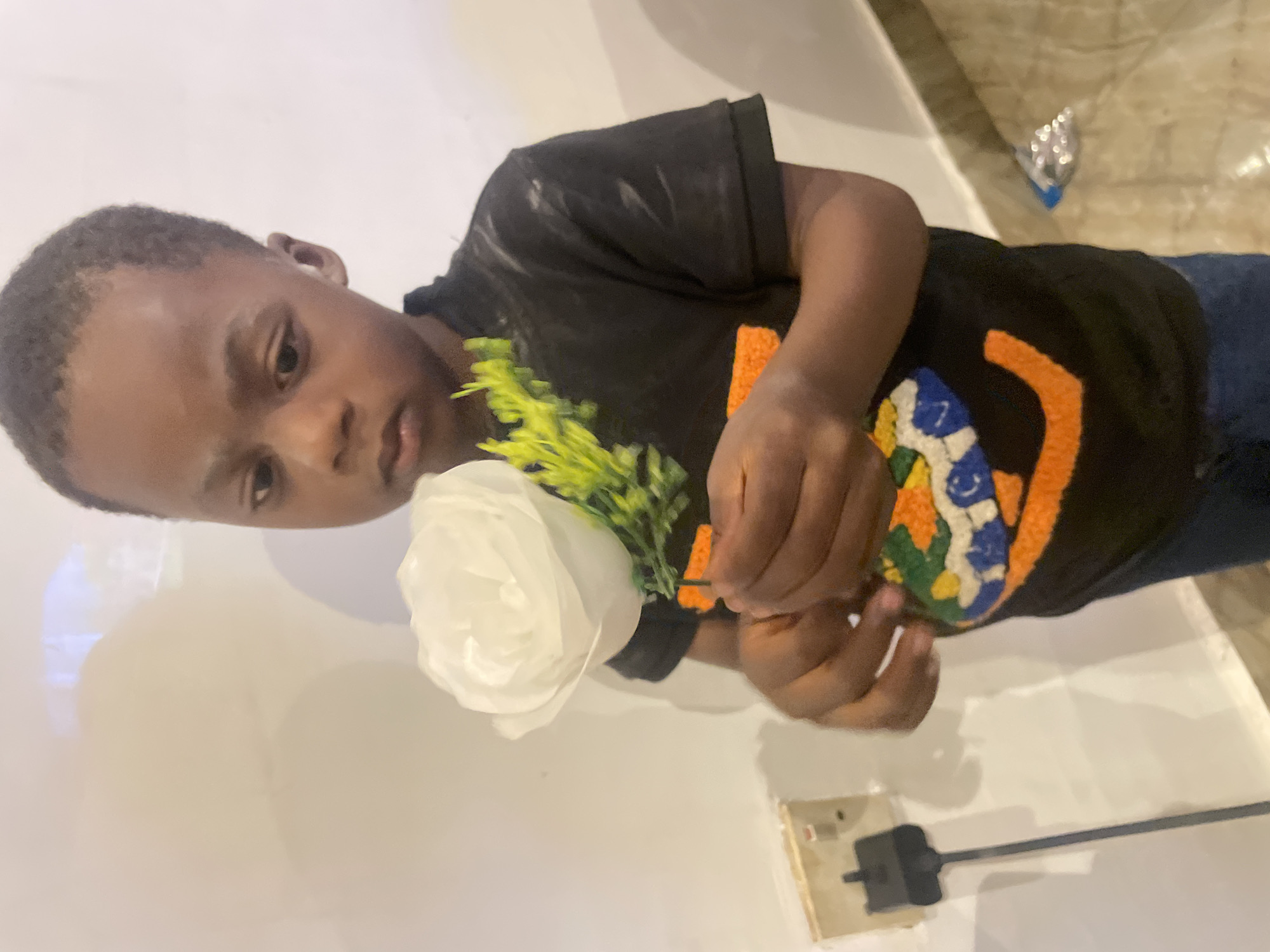 A young boy is holding a white rose. The boy is wearing a black shirt with an orange and yellow design on the front. He is standing inside. A power outlet is visible on the wall behind him. The location in the city of Jos, Nigeria is unknown.
