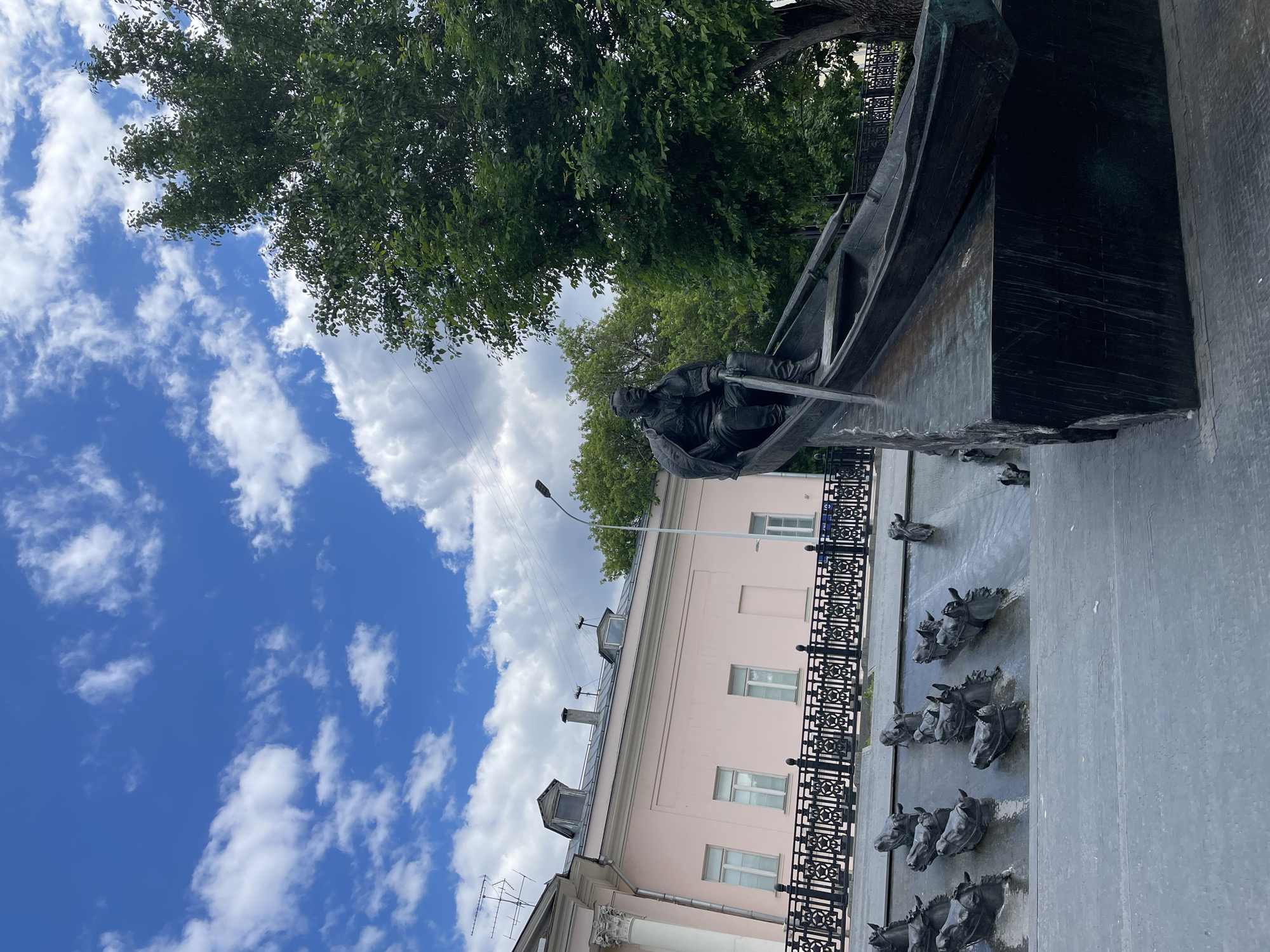 This image depicts a grand bronze monument located outdoors on a clear, sunny day, likely in Moscow, Russia. The central feature is a large sculpture of a figure, appearing to be a man, seated within a boat and holding oars. The boat is rendered in dark bronze with discernible details.
Below the boat, the monument incorporates a decorative fountain. A textured, dark wall serves as the backdrop for multiple stylized bronze horse heads, arranged in rows, which appear to function as water spouts, though no active water flow is visible. A black, ornate metal railing runs horizontally across the lower midground, separating the monument from the foreground.
In the background, a light pink, multi-story building with several white-framed windows is visible. Its roof features various dormer windows and chimneys. A tall street lamp stands near the building. Lush green trees with dense foliage frame the upper and left sides of the image, providing a natural contrast to the urban structures and the bright blue sky dotted with scattered white, fluffy clouds. The overall scene suggests a public park or square on a pleasant, bright day. No visible text is present in the image.