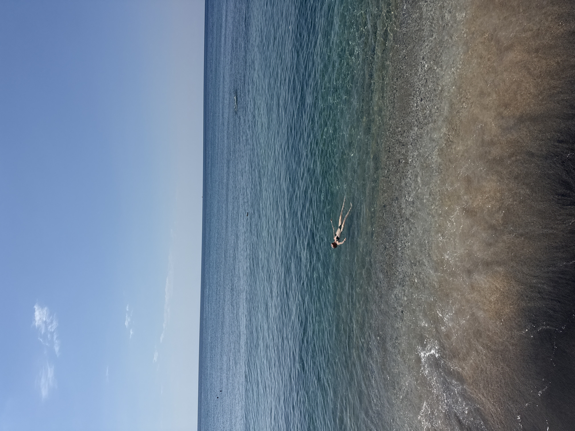 The image depicts a serene scene of a person floating on their back in the clear blue waters of Abang, Indonesia. The individual, wearing a dark bikini, is positioned horizontally in the water, with their arms extended slightly. The water is calm, with gentle ripples on the surface, and the seabed is visible through the transparent water, revealing a mixture of sand and pebbles. The background is dominated by the vast expanse of the ocean meeting a clear blue sky, with a few wispy clouds adding a touch of detail. The shoreline, composed of dark sand and pebbles, gradually transitions into the shallow, crystal-clear water. The overall atmosphere is one of tranquility and relaxation, capturing a moment of peaceful immersion in nature.