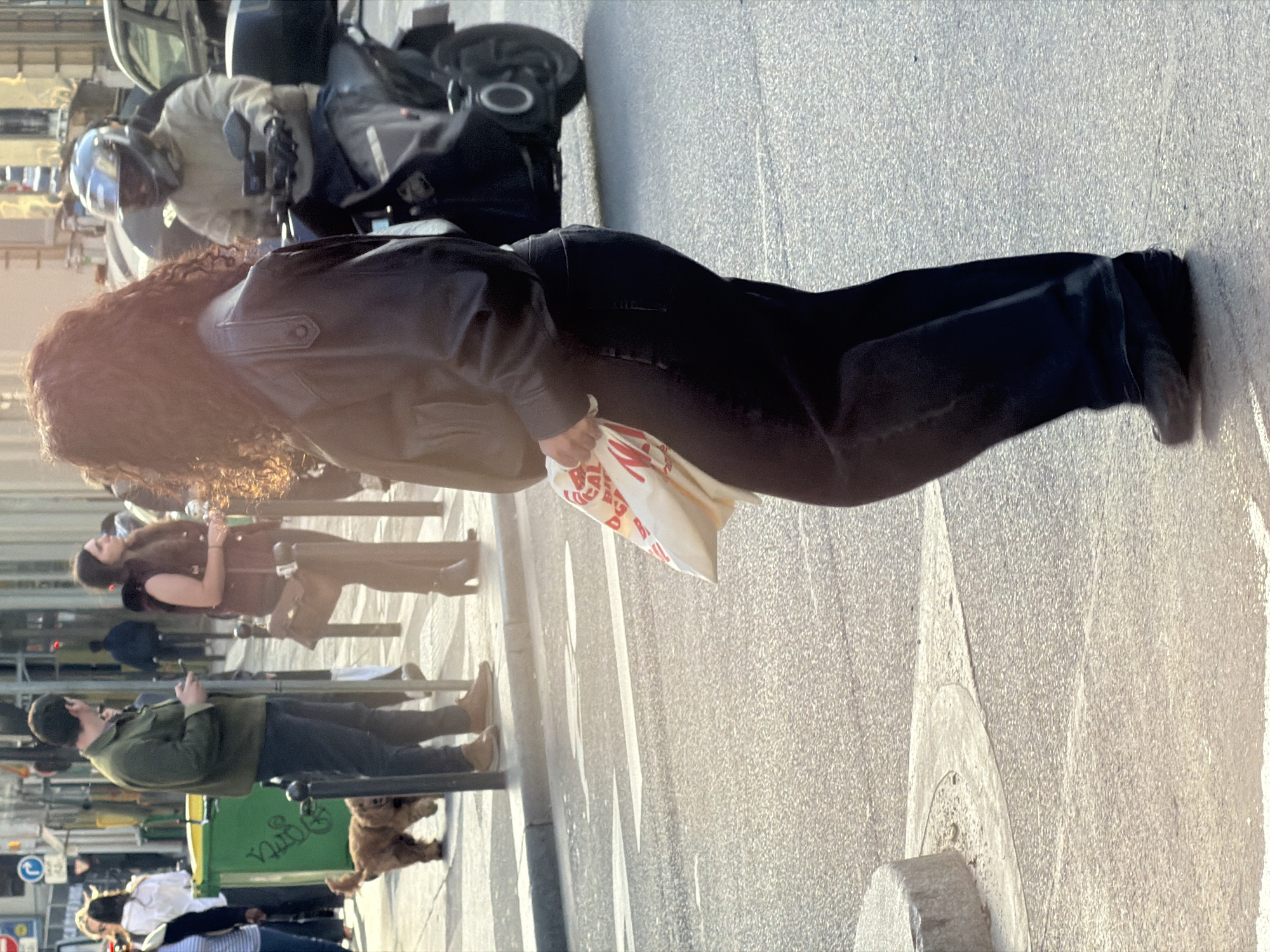 This image depicts a bustling street scene in Paris, France, captured on a clear, sunny day, likely during the late afternoon due to the warm, golden light and visible shadows. The central figure is a person with long, curly, reddish-brown hair, viewed from behind and slightly to the side. They are dressed in a black leather jacket and black pants, carrying a white shopping bag with repeating red text that is not clearly legible.
In the immediate background, a person wearing a light-colored jacket and a dark helmet is seated on a scooter or motorcycle. Further along the sidewalk, other pedestrians are visible: a woman in a brown top, a man in a green jacket accompanied by a small, light brown dog on a leash, and another individual in a white shirt.
The setting is an urban street featuring a paved road, a sidewalk with a curb, and visible white crosswalk lines. Various street furniture includes poles and a distinctive green trash bin with white text, partially reading "Paris." Buildings line the background, and a blue circular traffic sign with a white arrow, characteristic of European cities, is also discernible. The overall atmosphere is that of a typical, lively city thoroughfare.